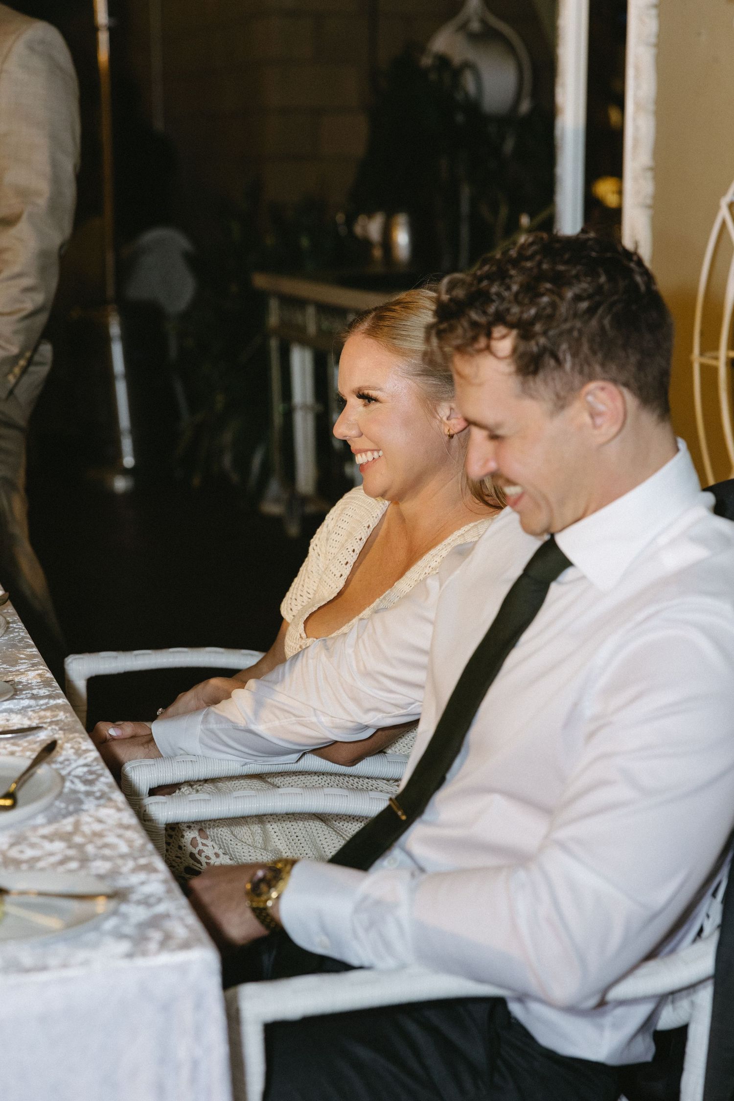Smiling bride and groom sit together at wedding reception table, woman in white dress, man in white shirt with black tie.