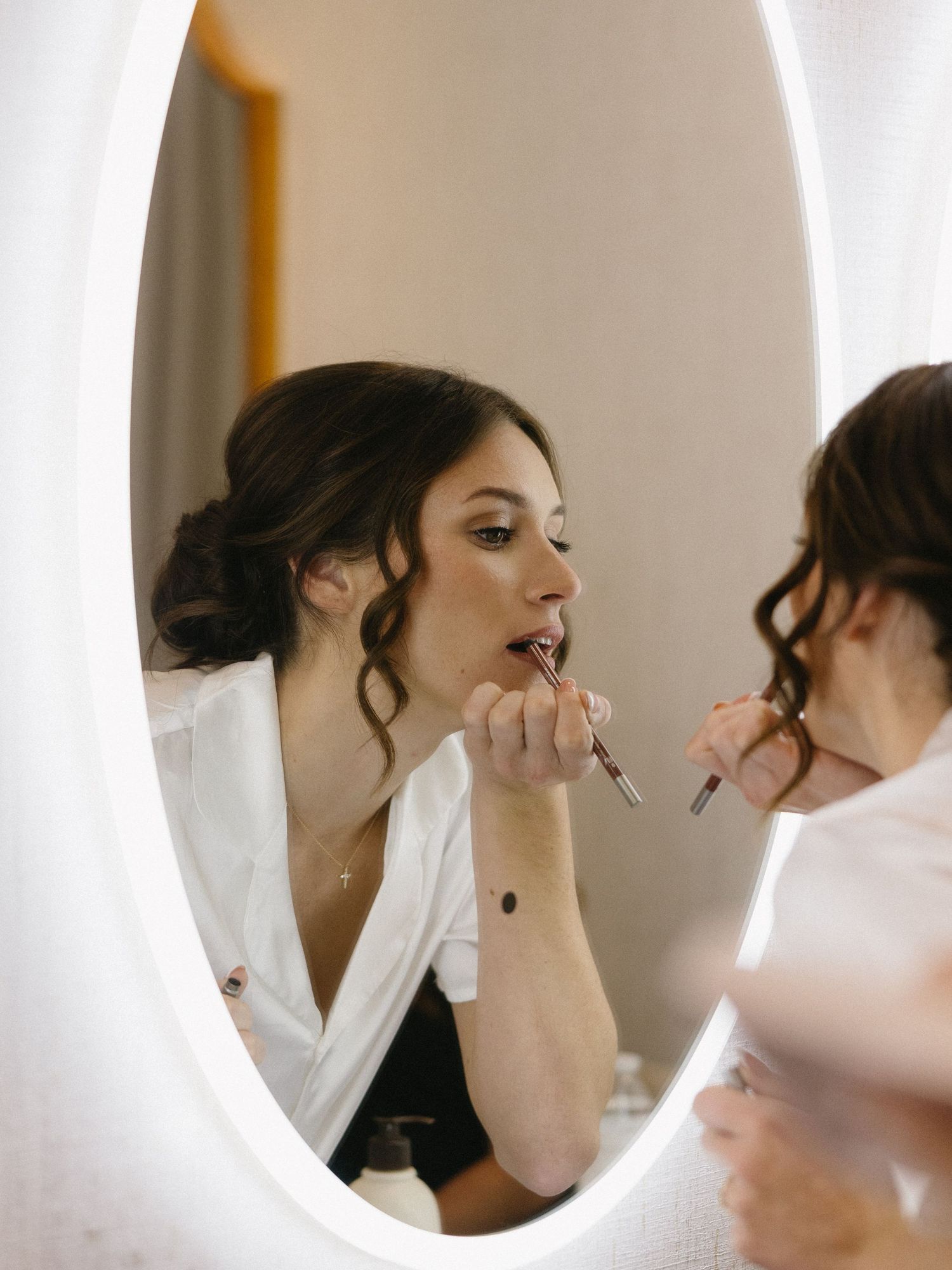 Bride getting lipstick applied by makeup artist, reflected in an oval lighted mirror while wearing a white robe.
