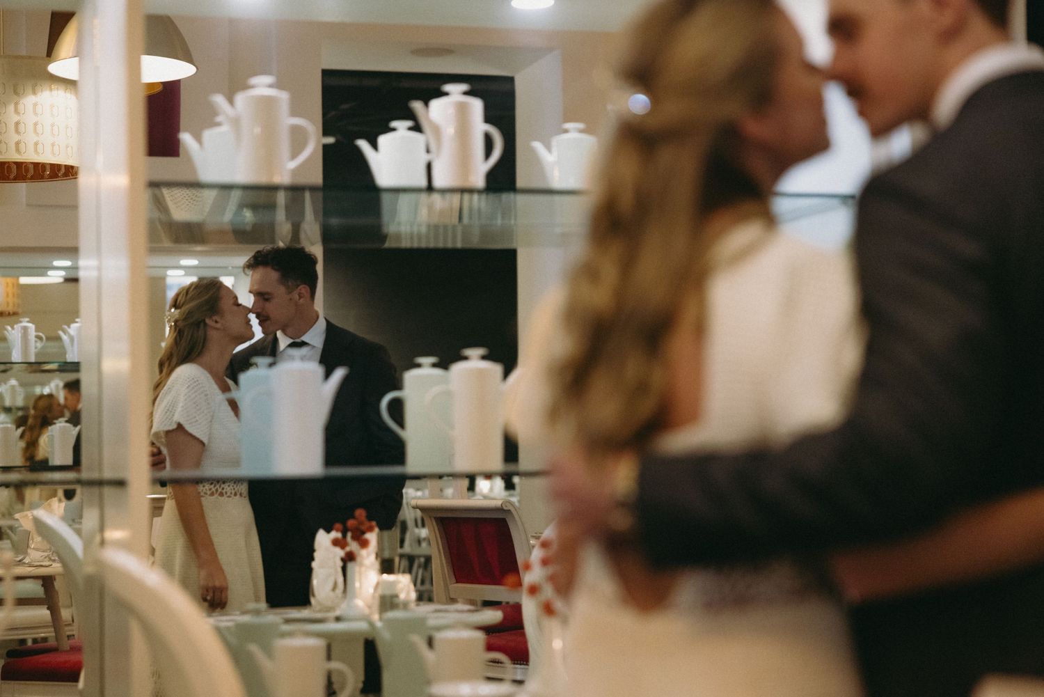 Bride and groom sharing a romantic kiss reflected in a mirror surrounded by elegant white china in a bridal shop.
