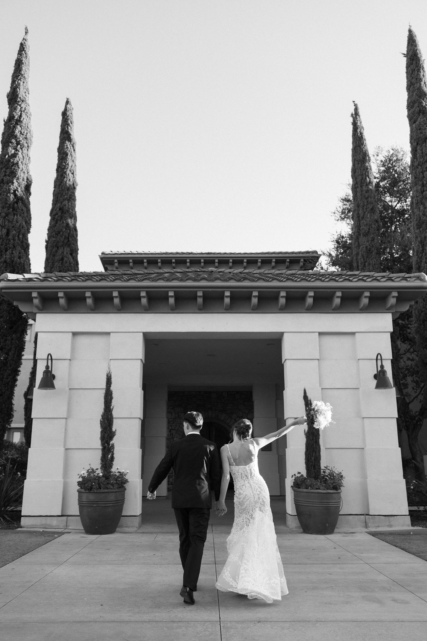 Wedding couple walking along modern white building with cypress trees.