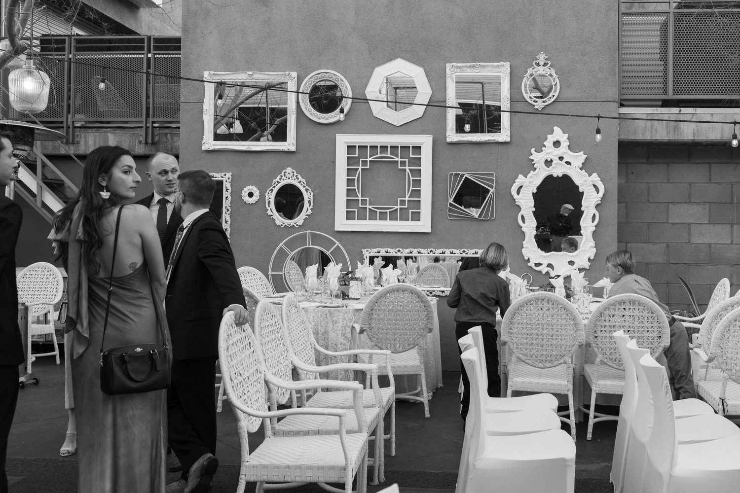 Black and white photo of elegantly dressed guests mingling at an outdoor event with decorative mirrors and wicker chairs.