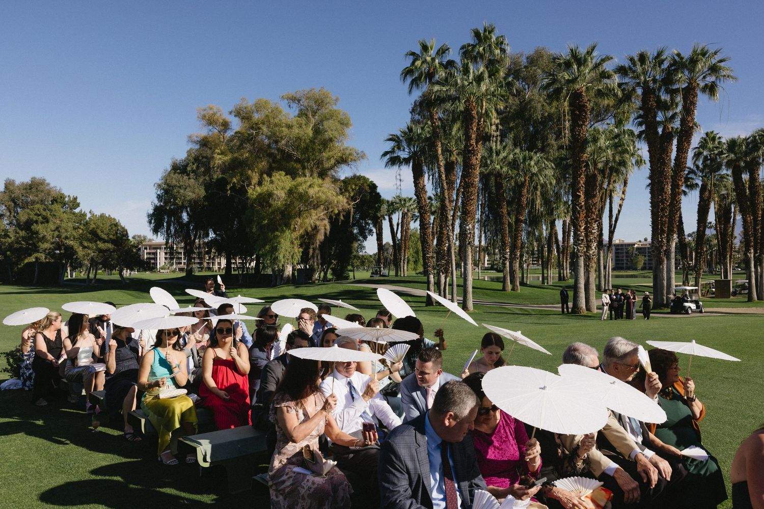 Outdoor wedding ceremony on lush golf course with guests holding white parasols, surrounded by tall palm trees.