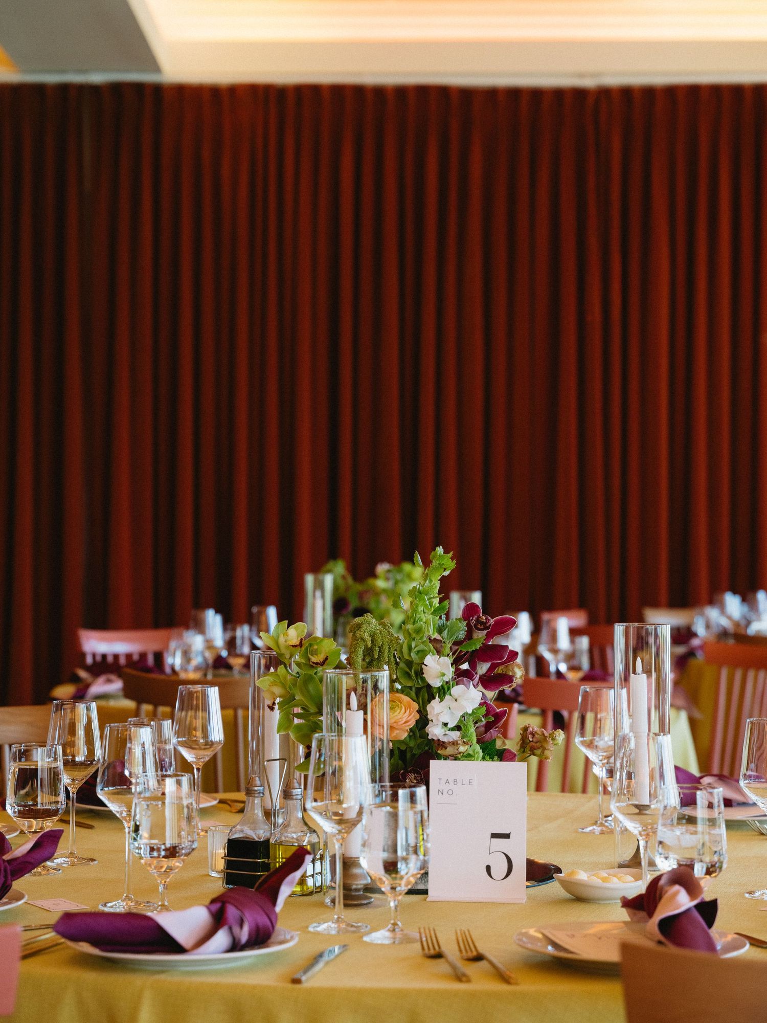 Elegant wedding reception table set with gold linens, purple napkins, floral centerpiece, and glassware against rich red curtains.