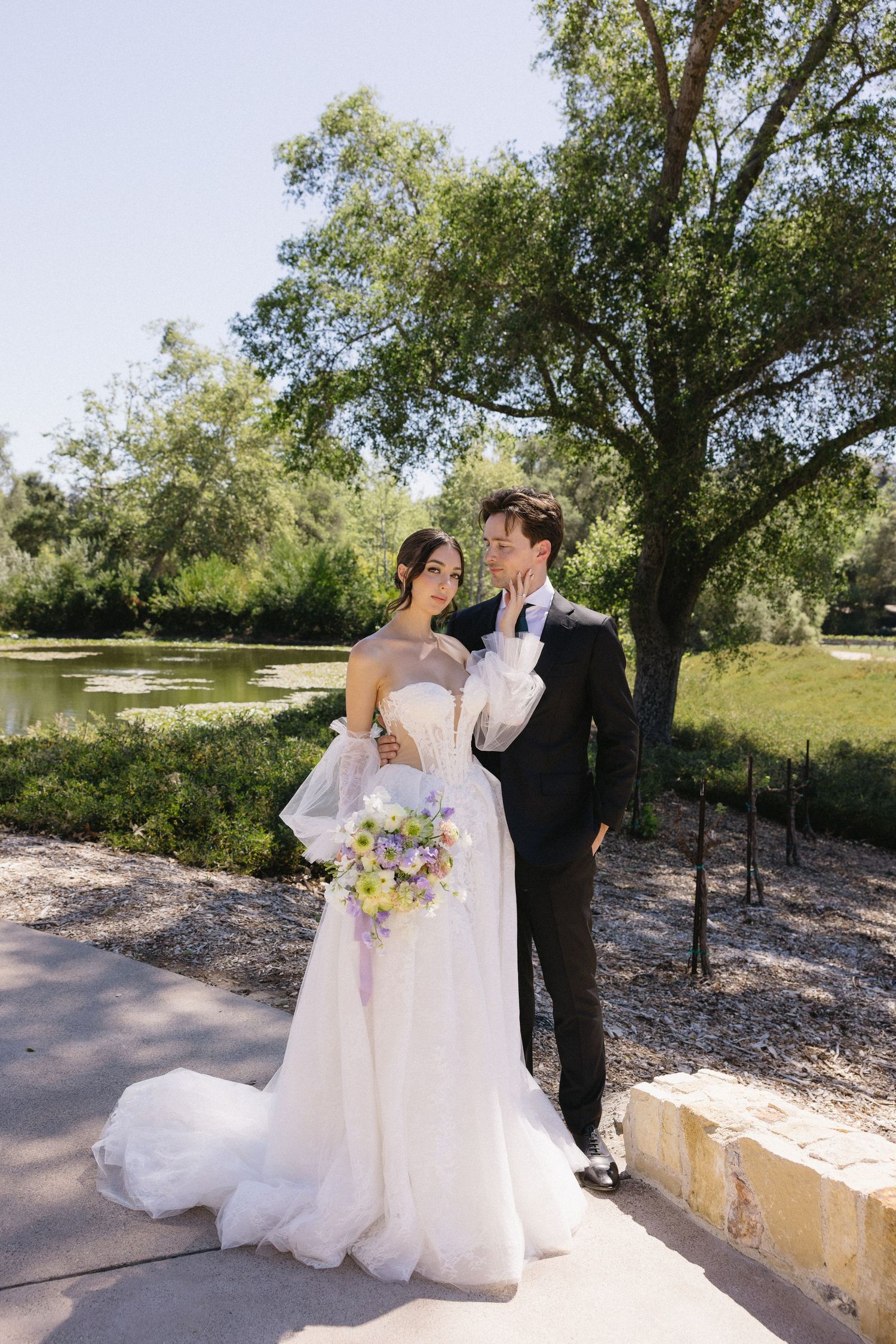 Bride in white ballgown holding floral bouquet poses with groom in black tuxedo outdoors near a serene pond.