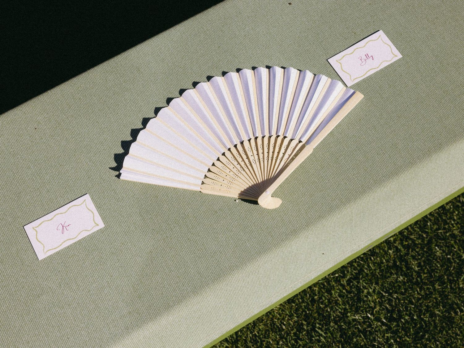 Elegant white paper hand fan with bamboo handle displayed beside monogram place cards on a green surface.