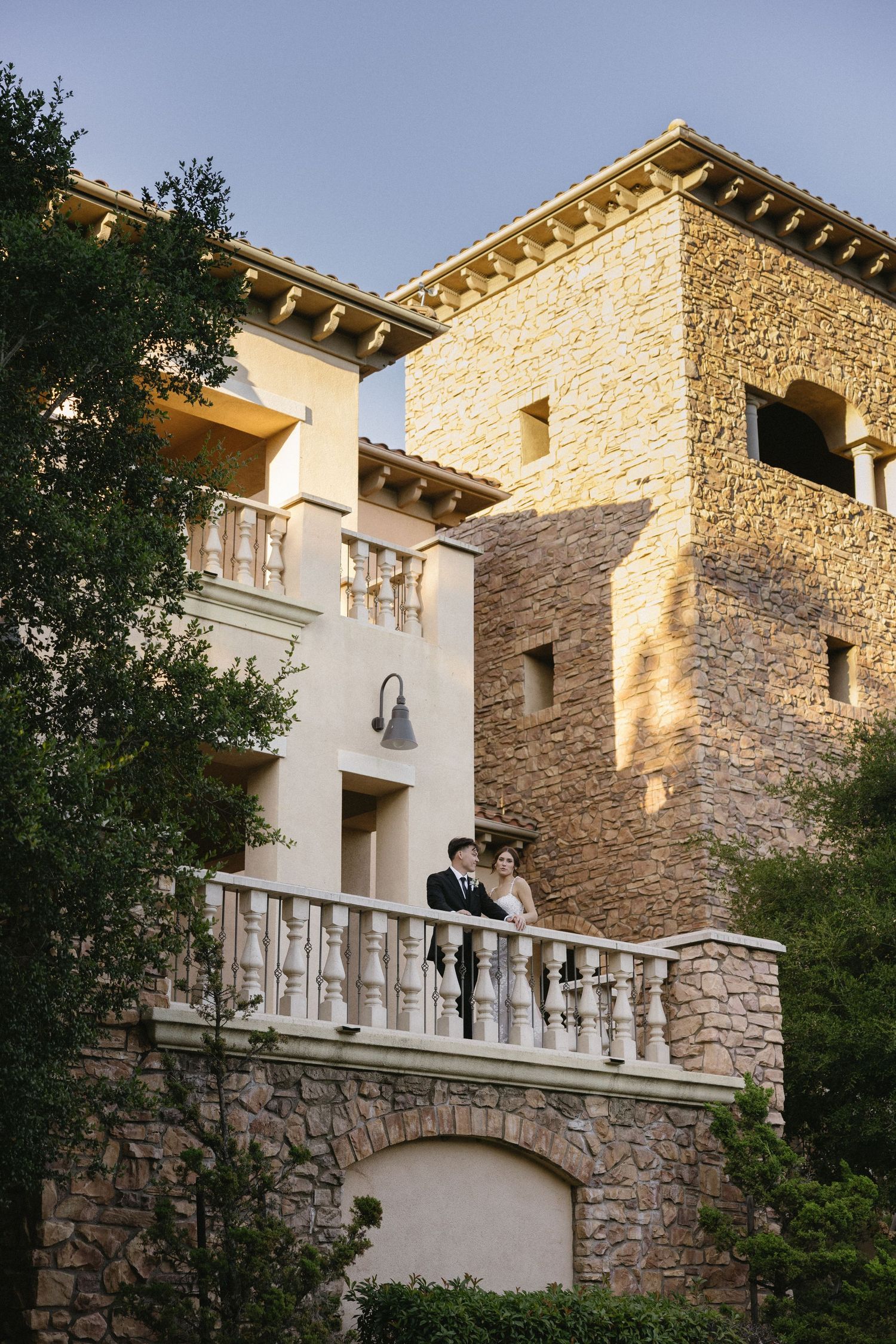 Bride and groom stand on elegant stone balcony of Mediterranean-style villa surrounded by lush trees at golden hour.