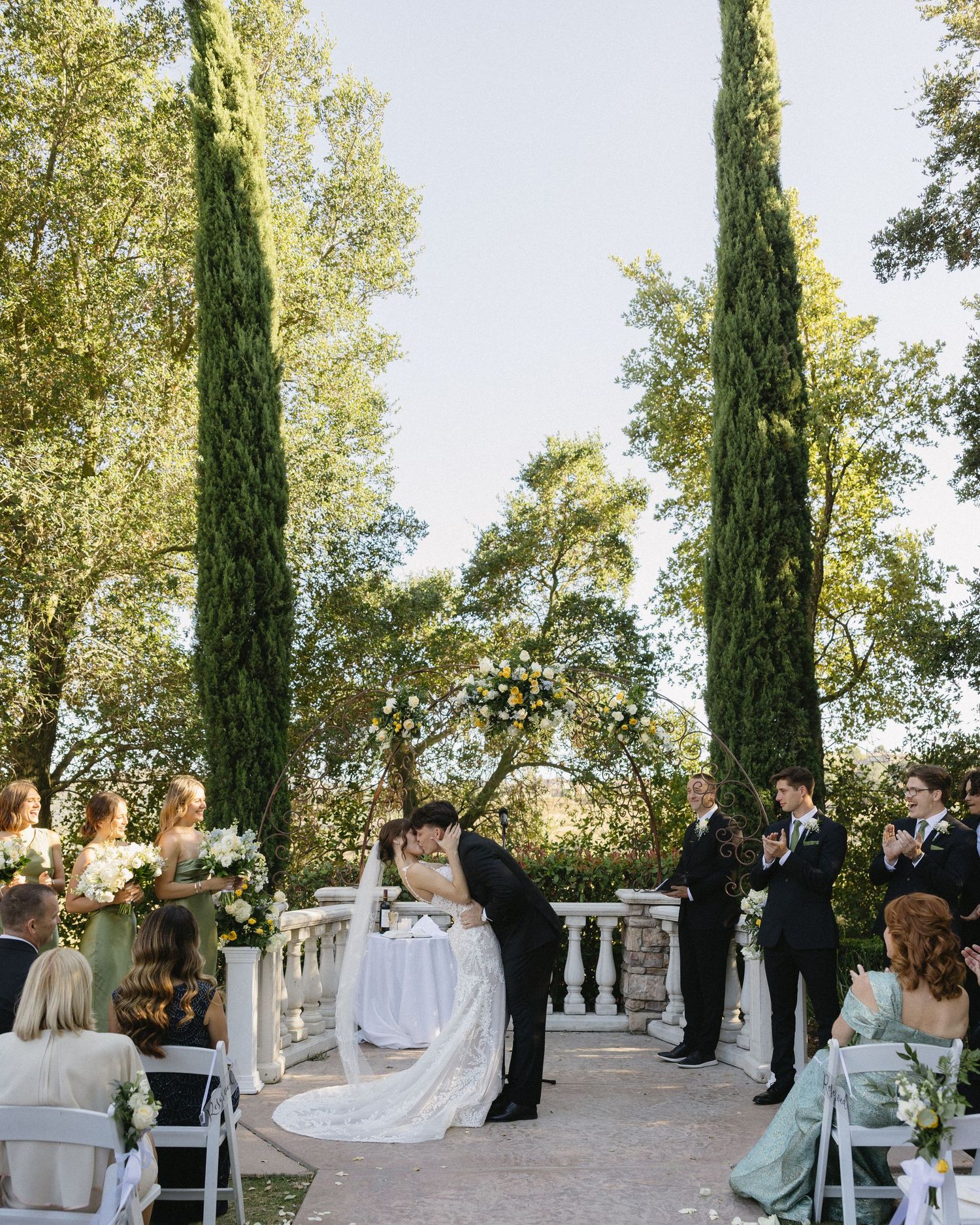 Couple shares first kiss at elegant outdoor wedding ceremony with cypress trees, white balustrade, and floral arch backdrop.