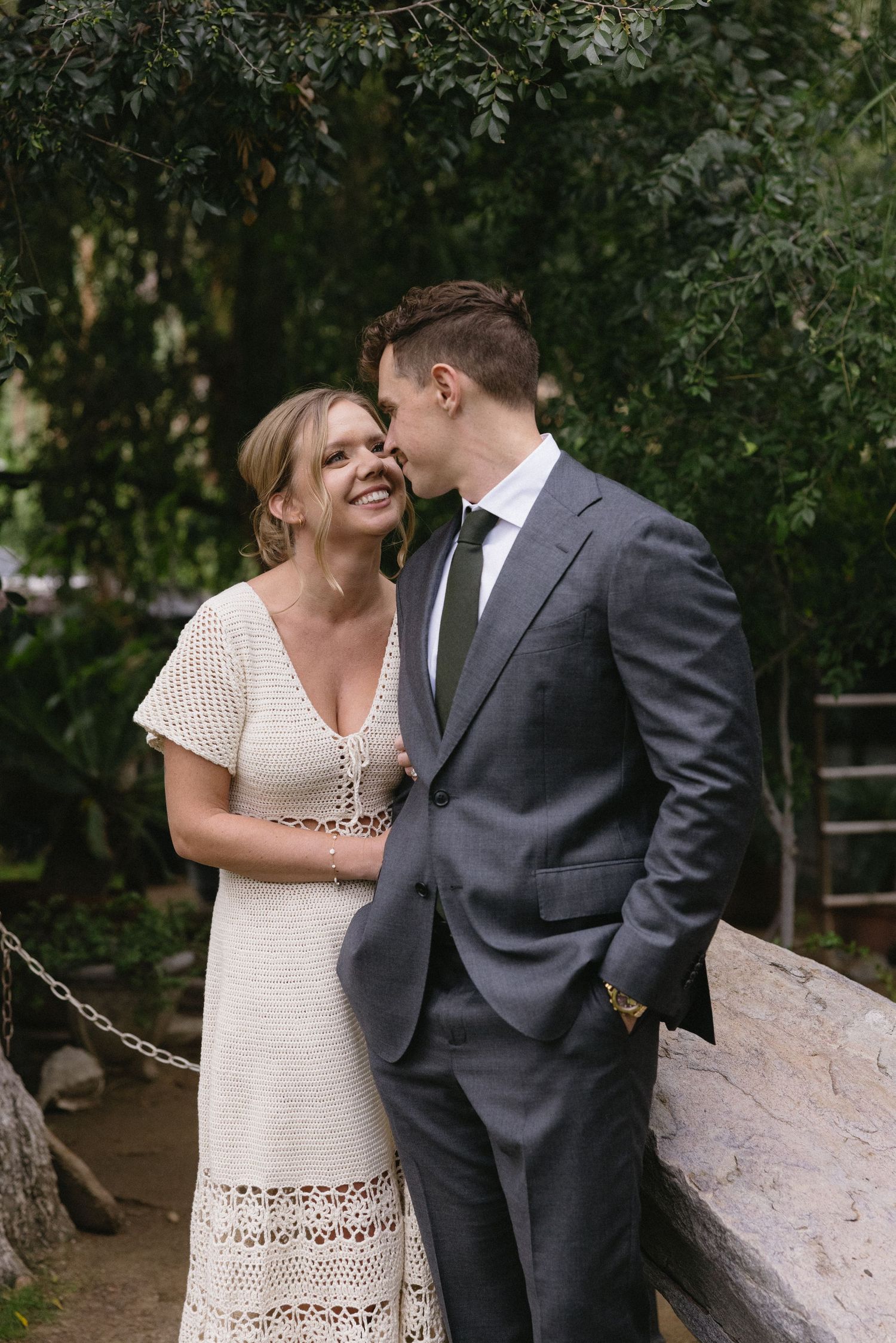 Couple sharing a romantic moment outdoors, woman in white crochet dress smiling at man in gray suit near stone wall.