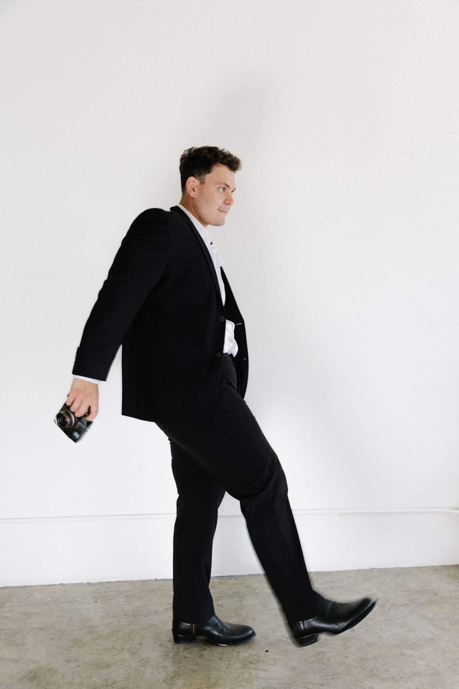 Man in black tuxedo walking confidently, holding a camera, against a white wall on a concrete floor.
