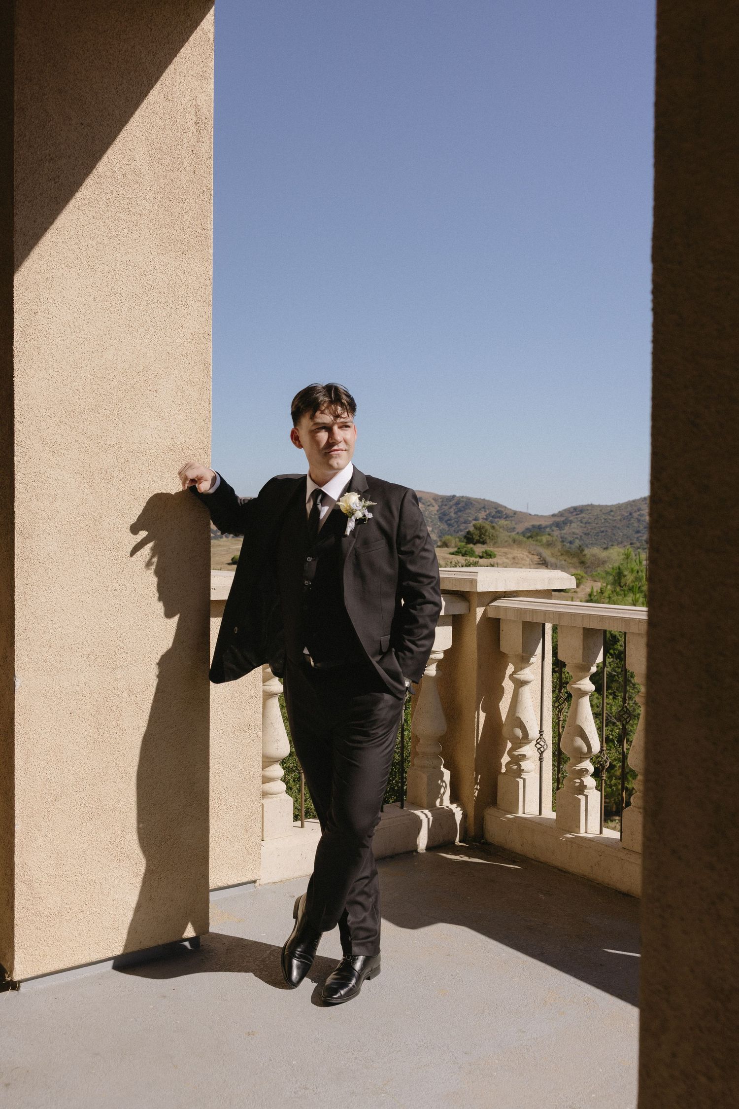 Groom in black suit with boutonniere poses on elegant balcony with mountain views and blue sky.