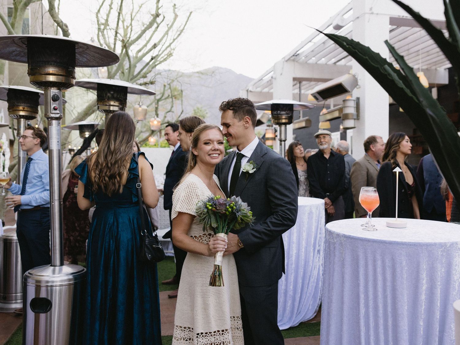 Bride and groom smile together at outdoor wedding reception with guests and patio heaters in background.