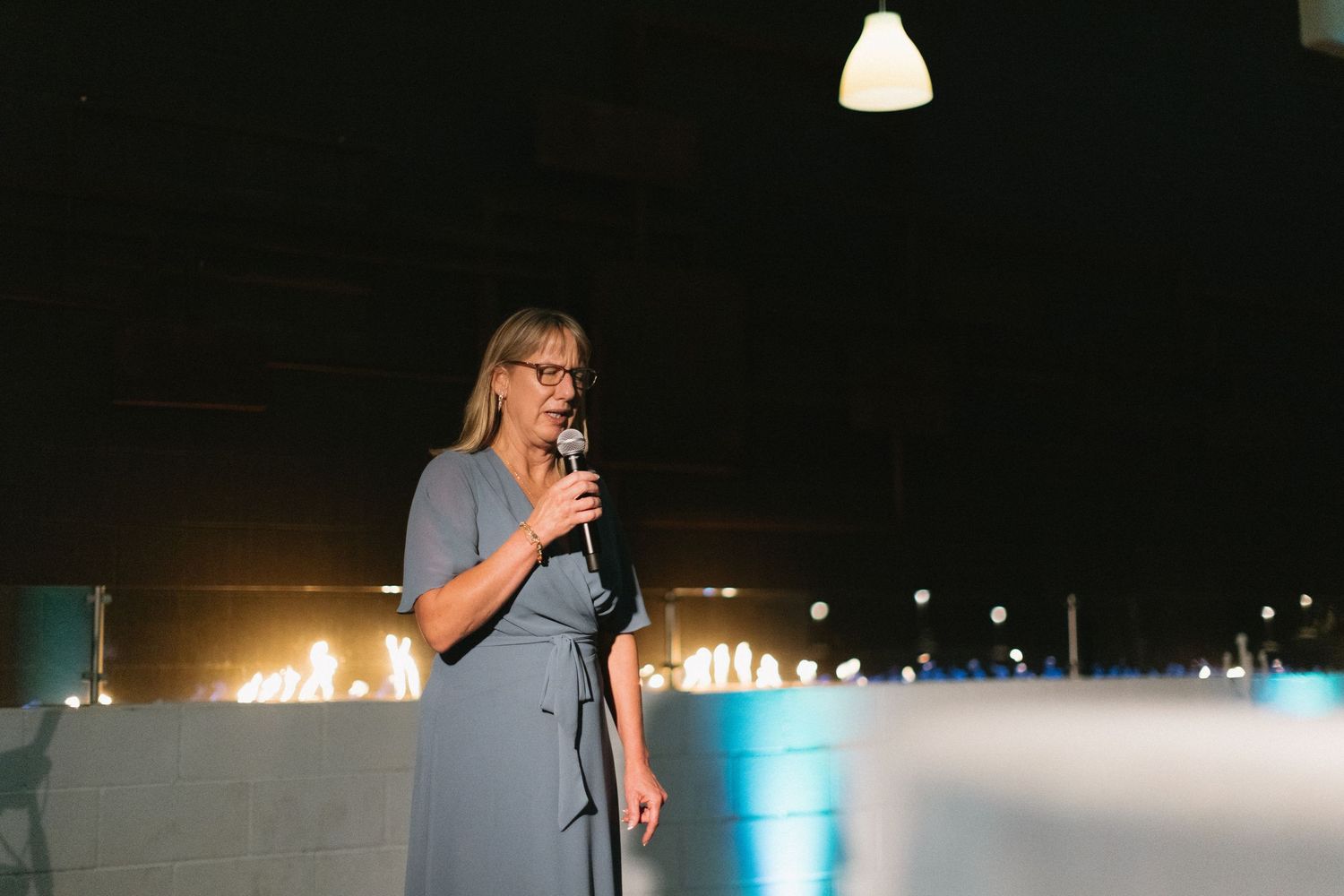 Woman in gray dress speaking into microphone at elegant evening event with candles in background.
