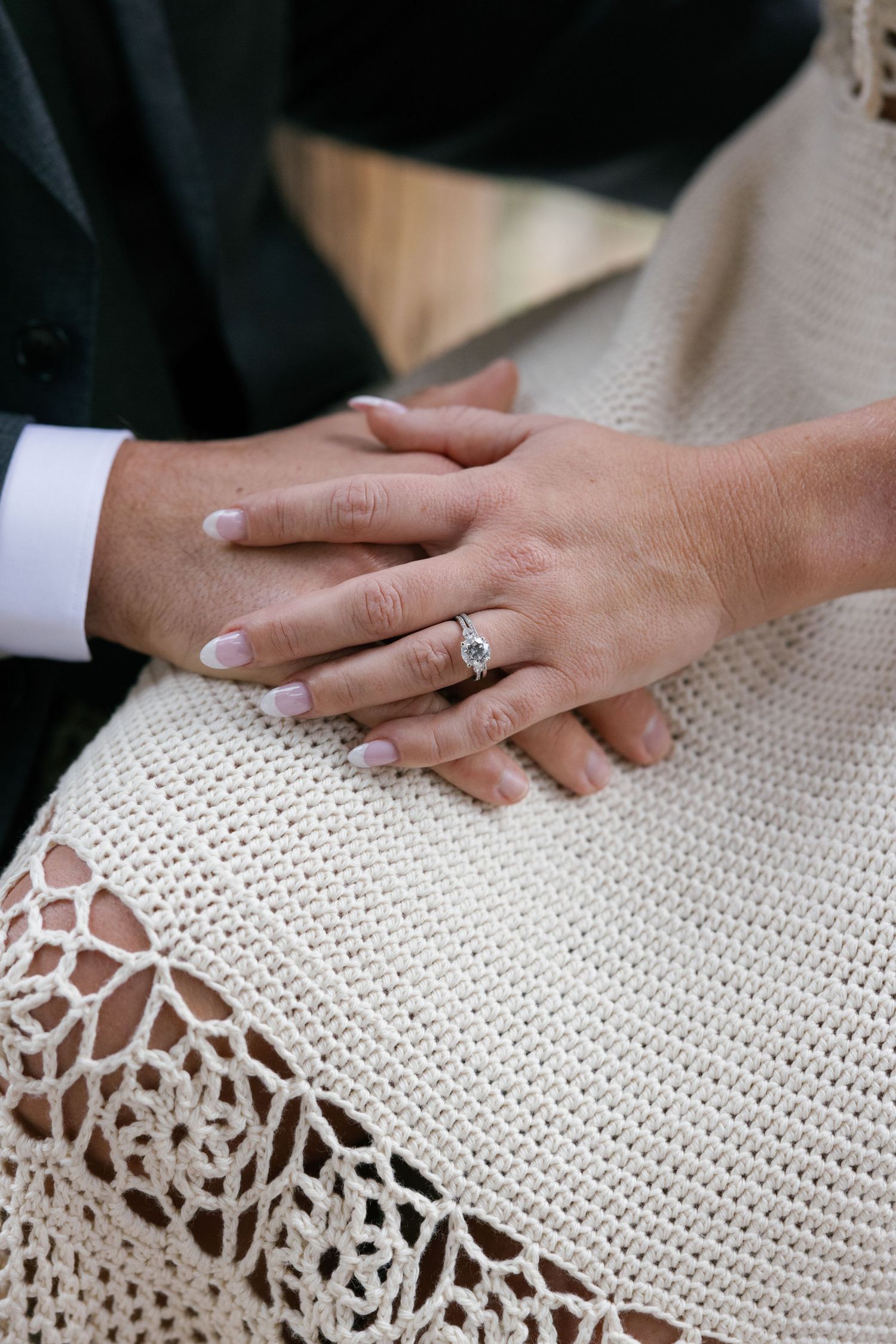Close-up of a couple's hands resting together, showcasing a diamond engagement ring on the bride's finger.