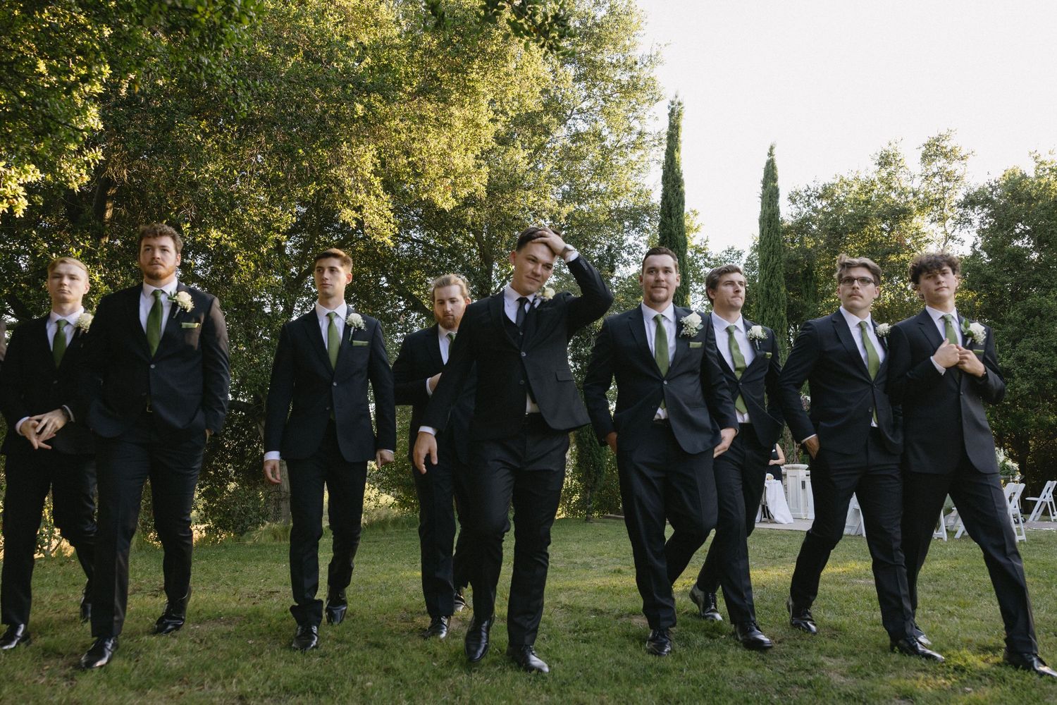 Groomsmen in black suits with green ties walk confidently outdoors at a wedding, surrounded by lush trees.