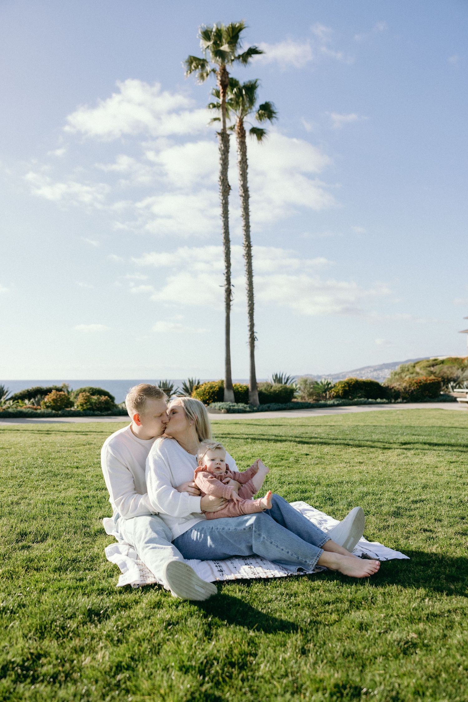A couple relaxes on a blanket in a grassy field with palm trees and mountains in the background.
