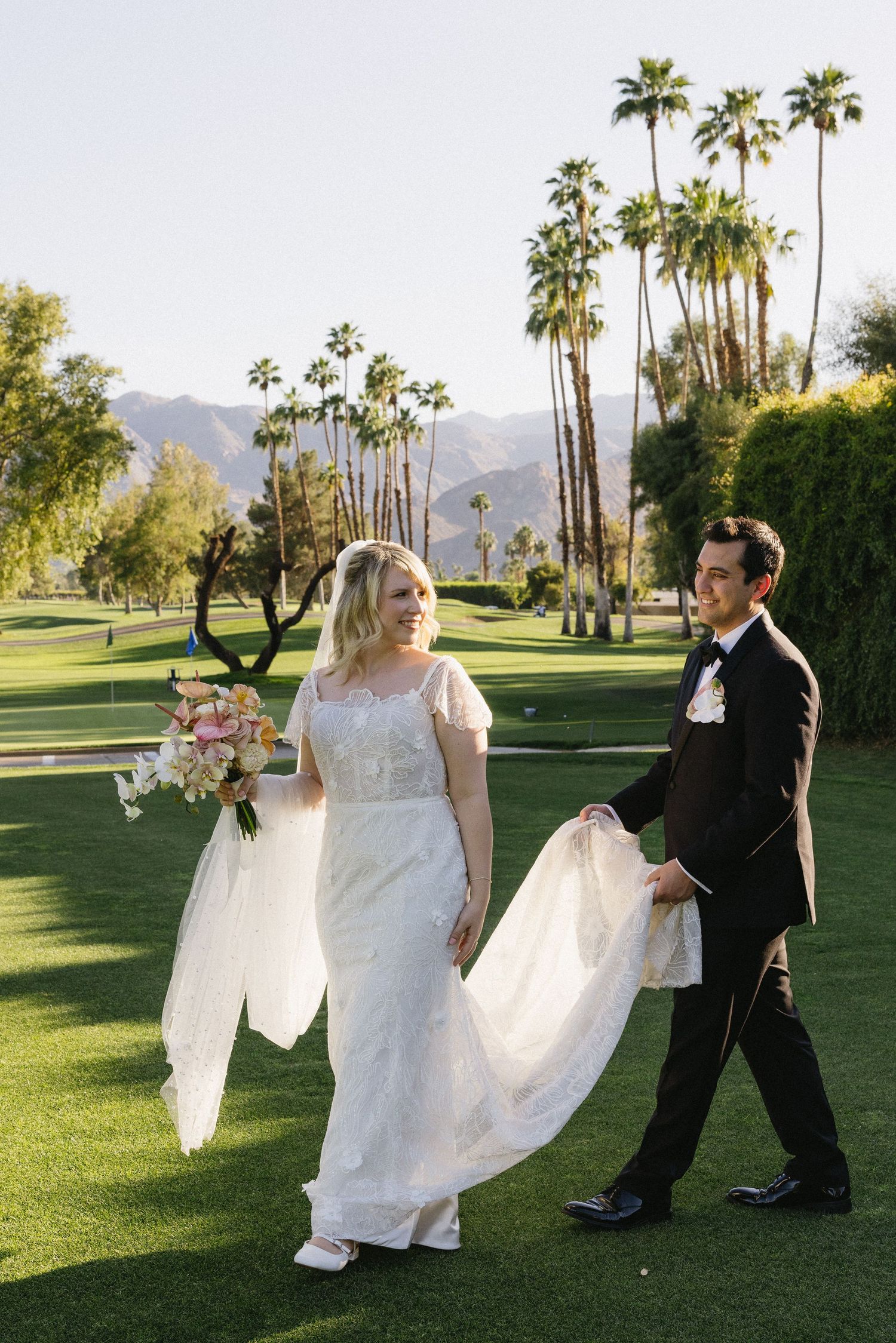 Bride and groom walk on lush green golf course with palm trees and mountains in Palm Springs wedding.