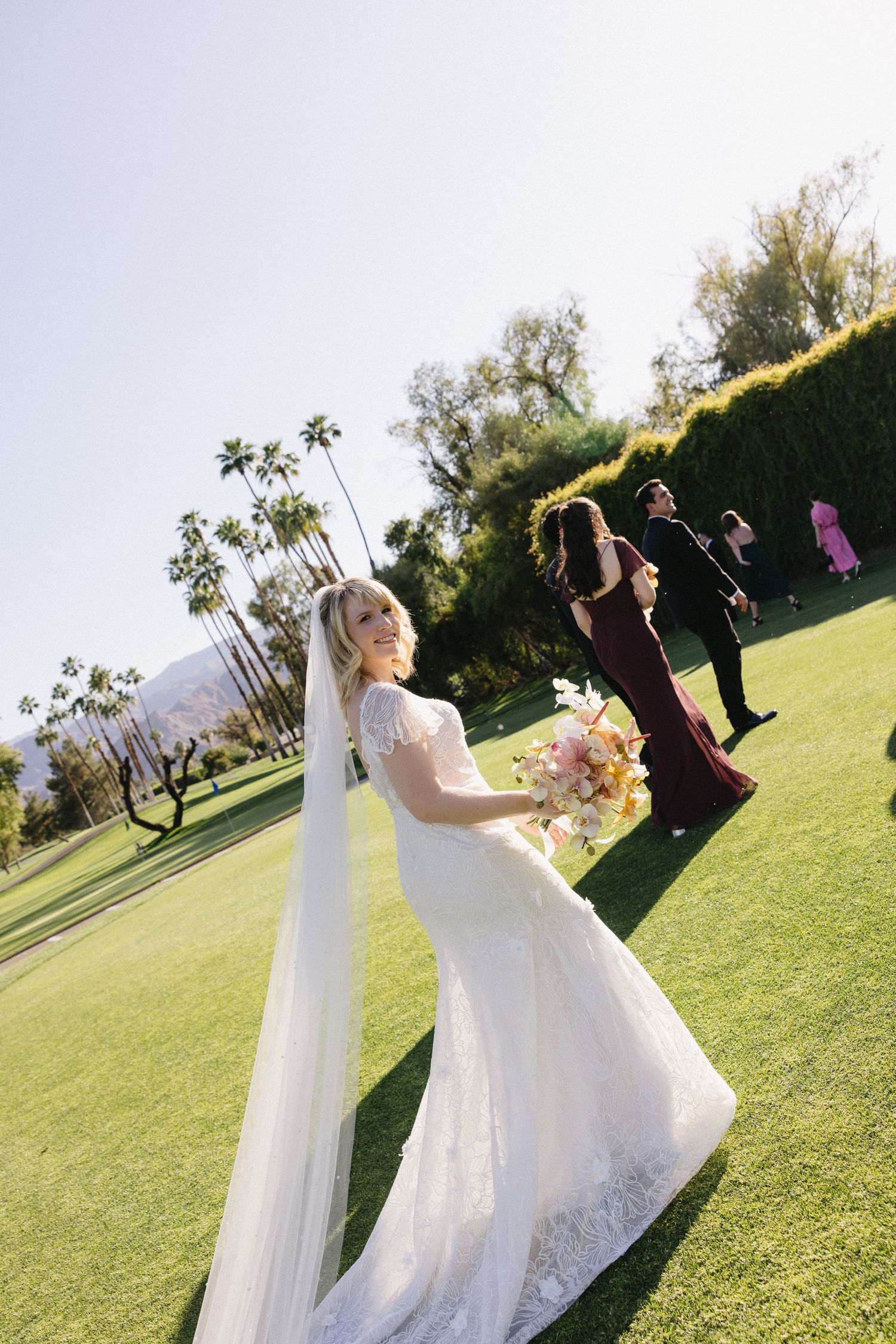 Bride in white lace gown and long veil holds colorful bouquet on green lawn with palm trees at outdoor wedding.