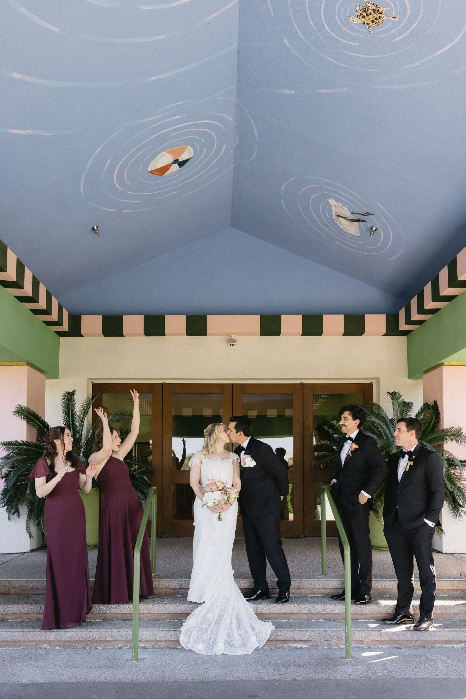 Bride and groom kissing on steps with wedding party in burgundy dresses and black tuxedos outside colorful venue.