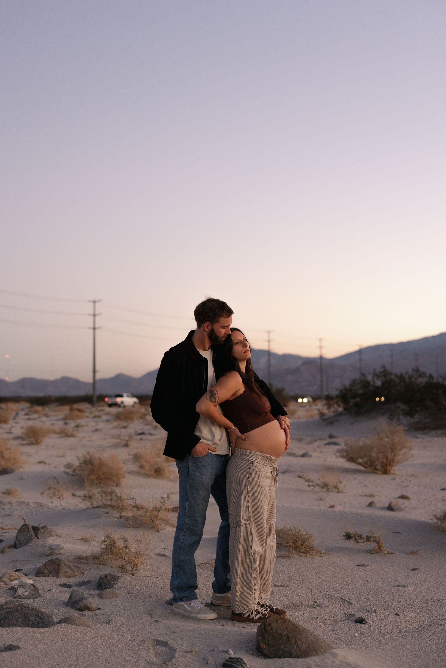 Couple standing together in desert landscape at dusk with mountains in background.