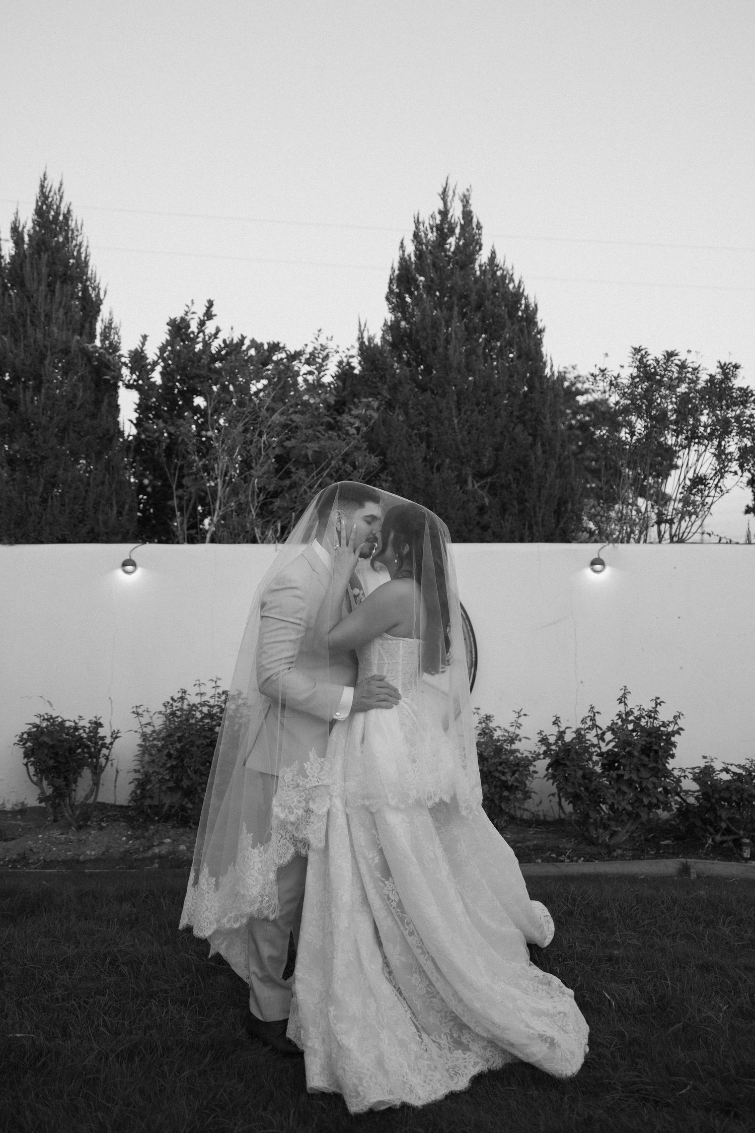 Couple in formal wedding attire twirling and dancing together in a garden setting at dusk.