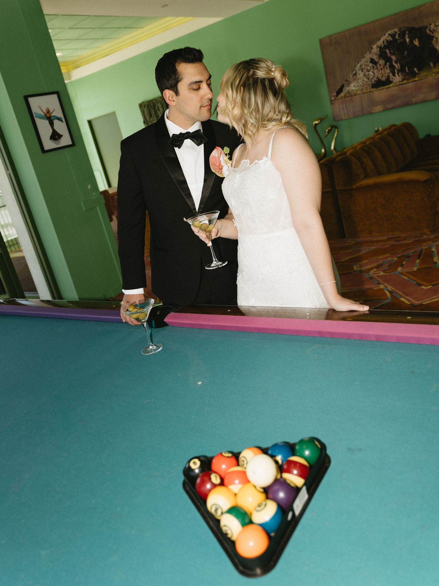 Bride and groom kissing over a pool table with racked billiard balls, holding martini glasses at their wedding reception.