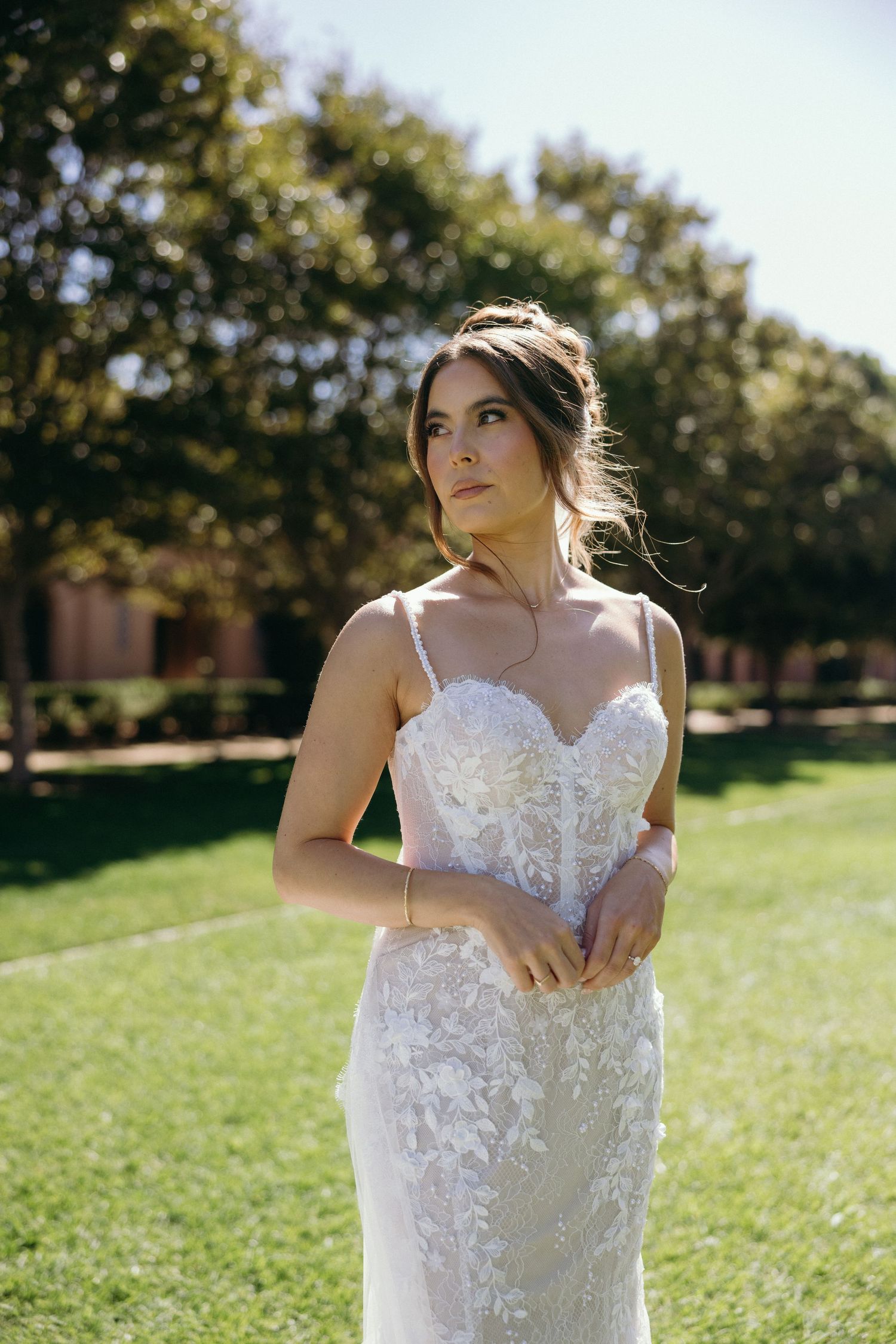 Bride in a lace spaghetti strap wedding gown standing on green grass with trees in the background.