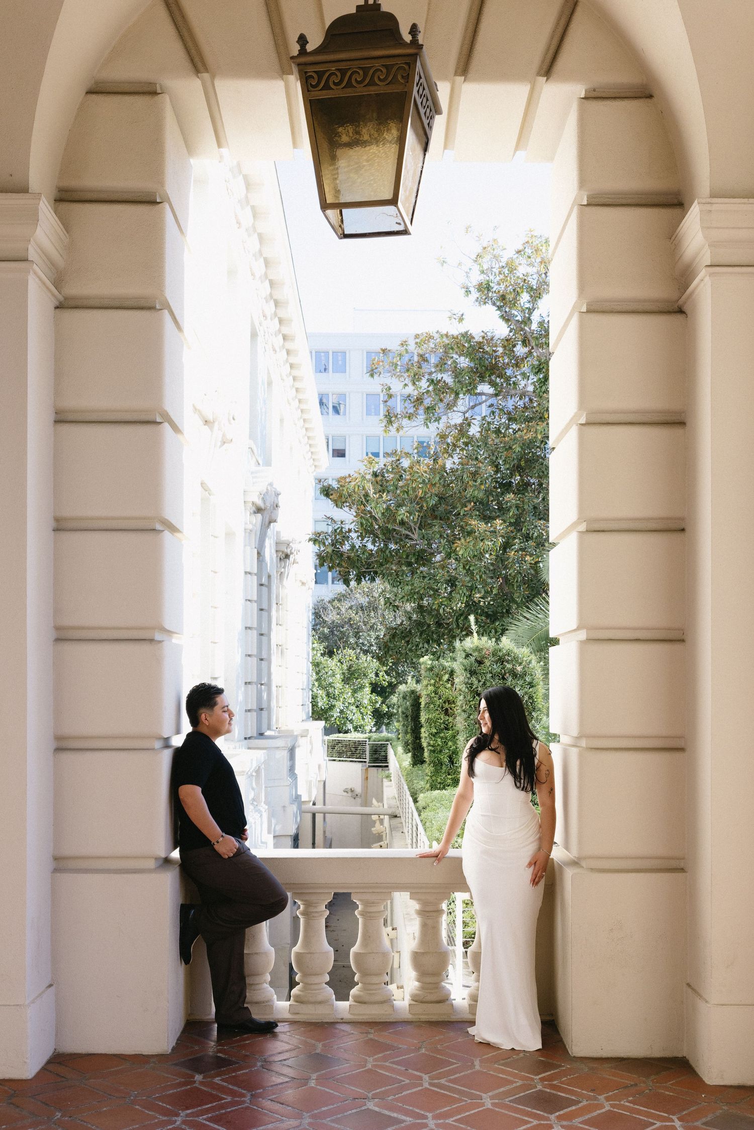 Couple in elegant attire standing under ornate arch with lantern, sharing a romantic moment at a classic venue.
