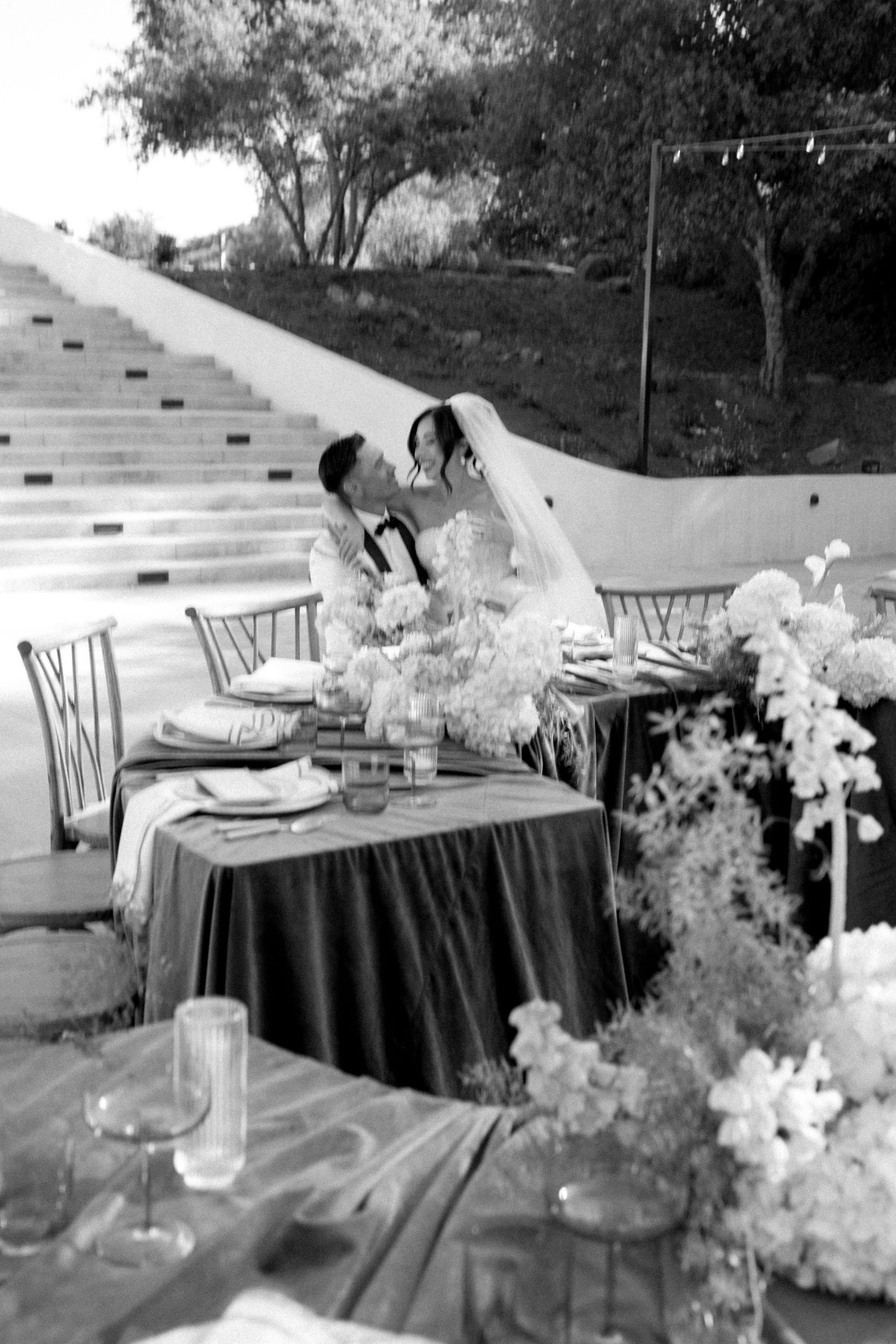 Black and white photo of a bride and groom sharing a kiss at their elegantly decorated outdoor wedding reception table.
