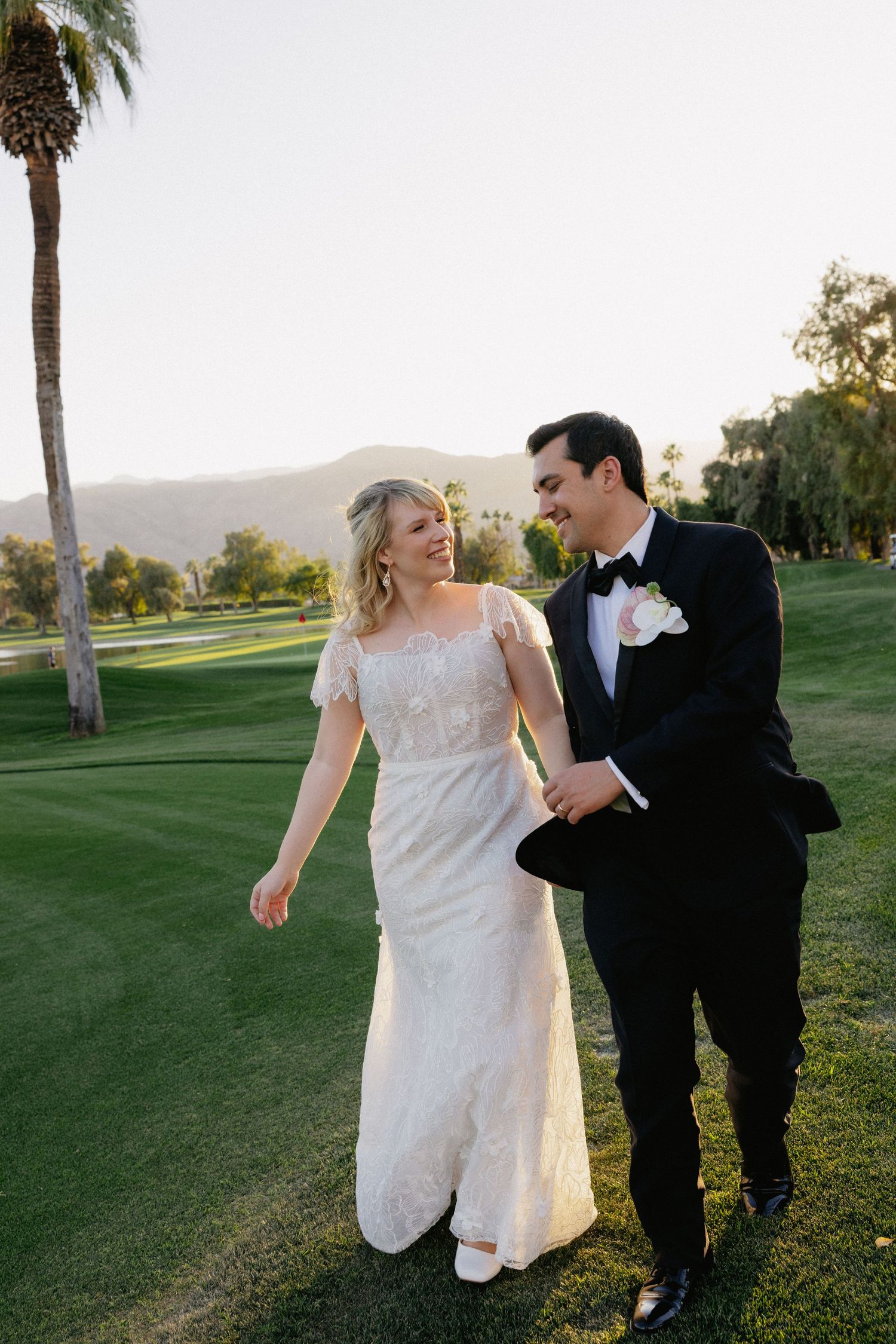 A bride and groom walk hand-in-hand on a lush green golf course with palm trees and mountains at sunset.