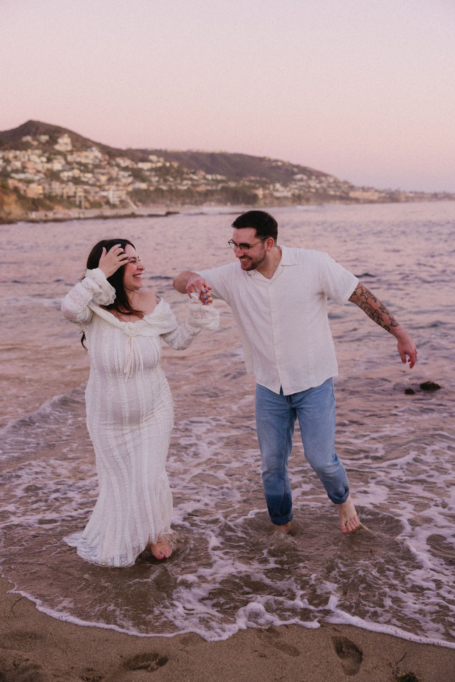 A playful couple splashes in the ocean waves at sunset near a rocky coastline.