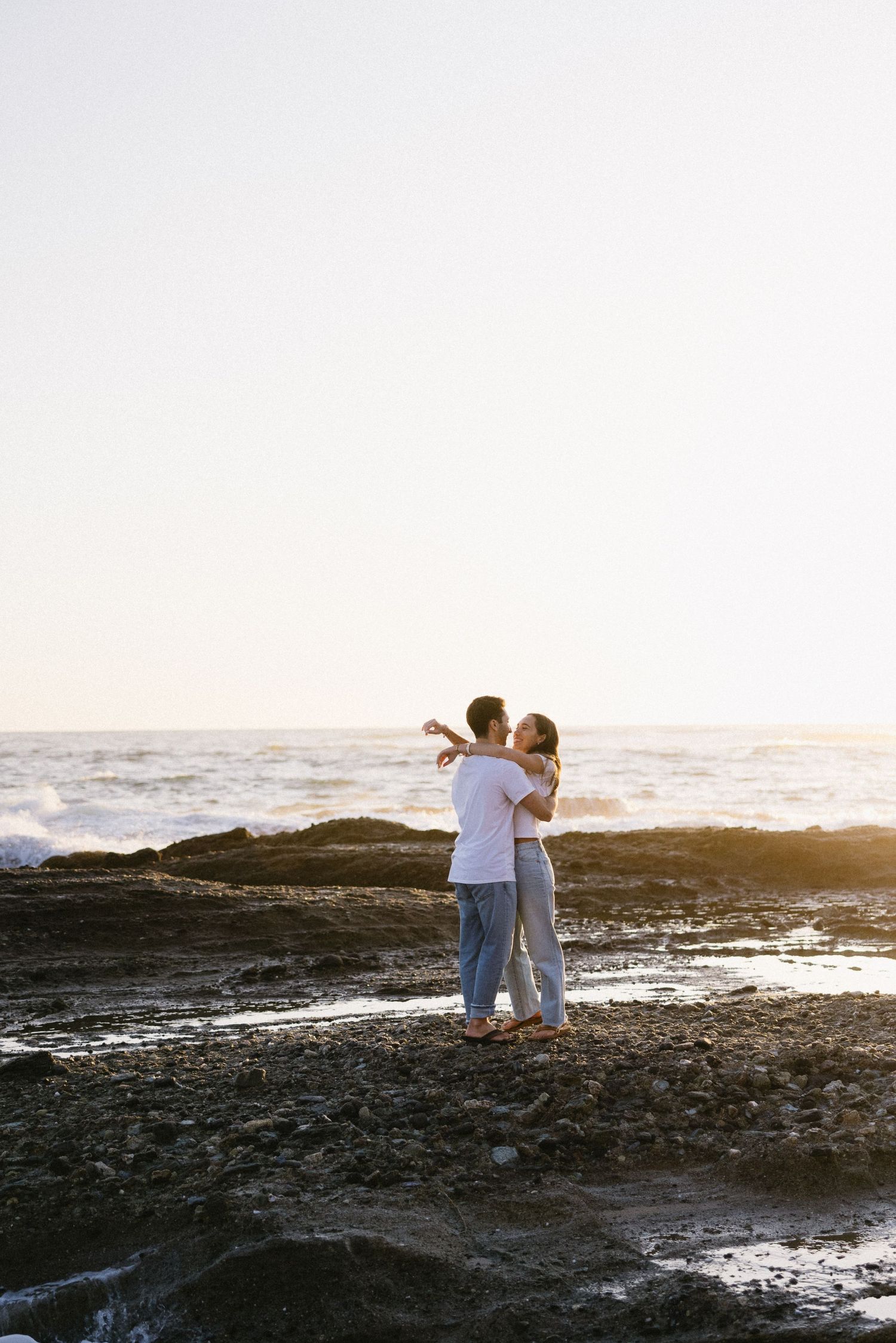 A couple sharing tender moments on rocky coastal shores during sunset.