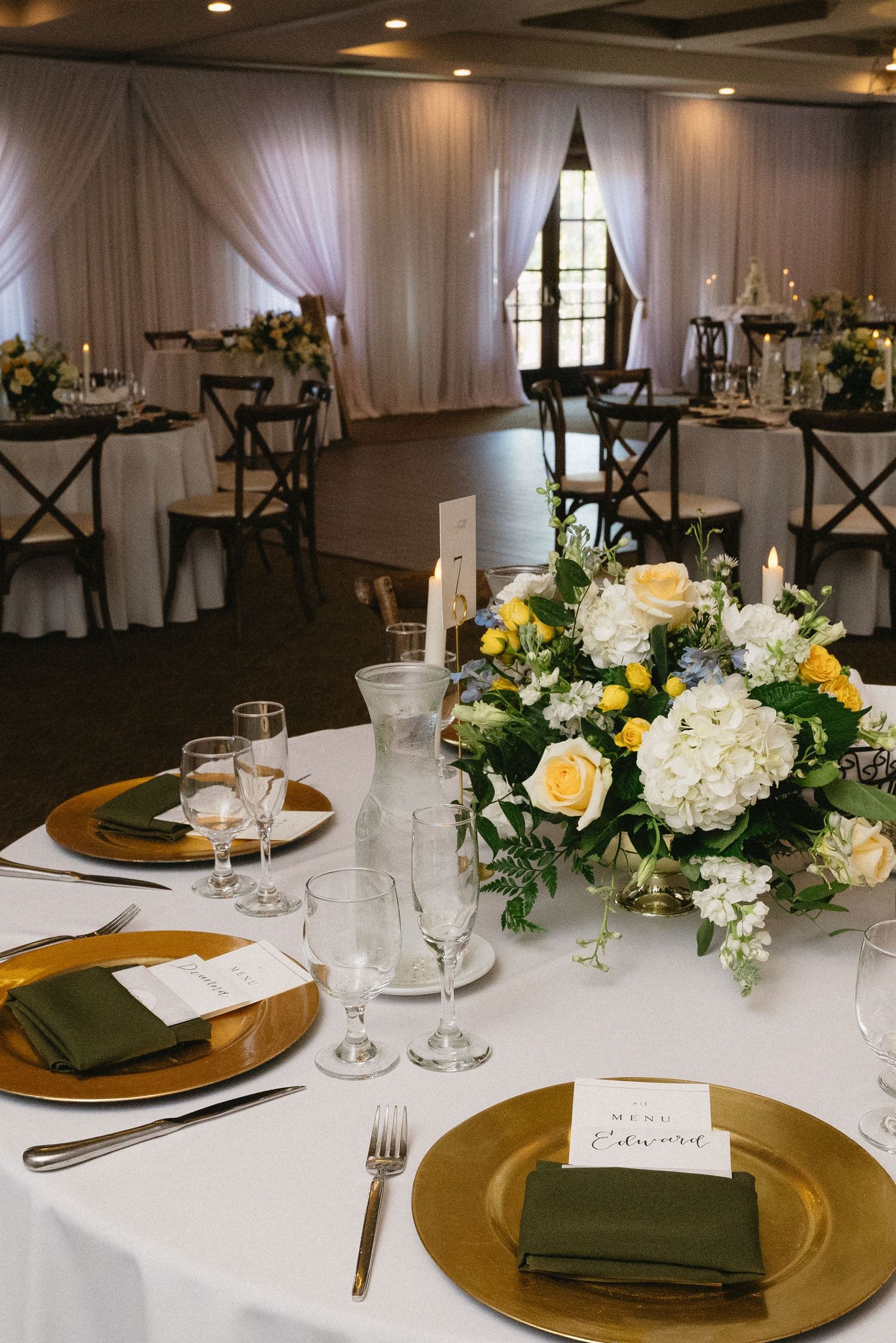 Elegant wedding reception table with gold chargers, green napkins, and yellow and white floral centerpiece in a draped ballroom.