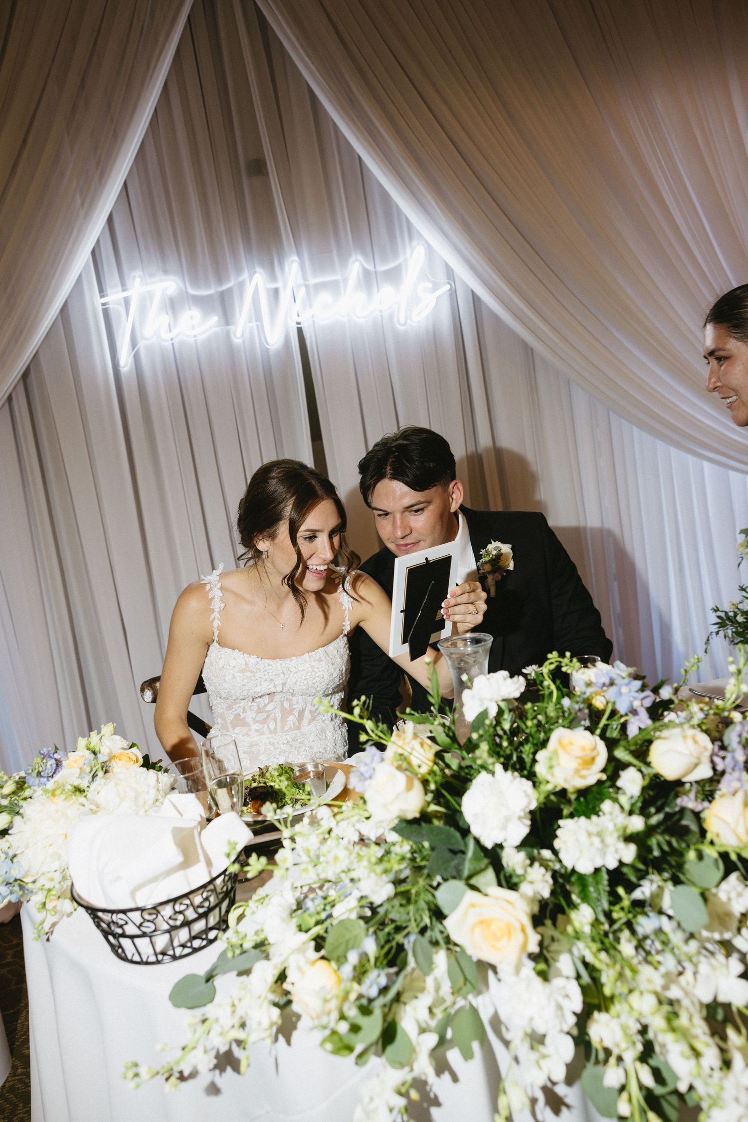 Bride and groom at sweetheart table with white floral arrangements and neon sign backdrop at wedding reception.