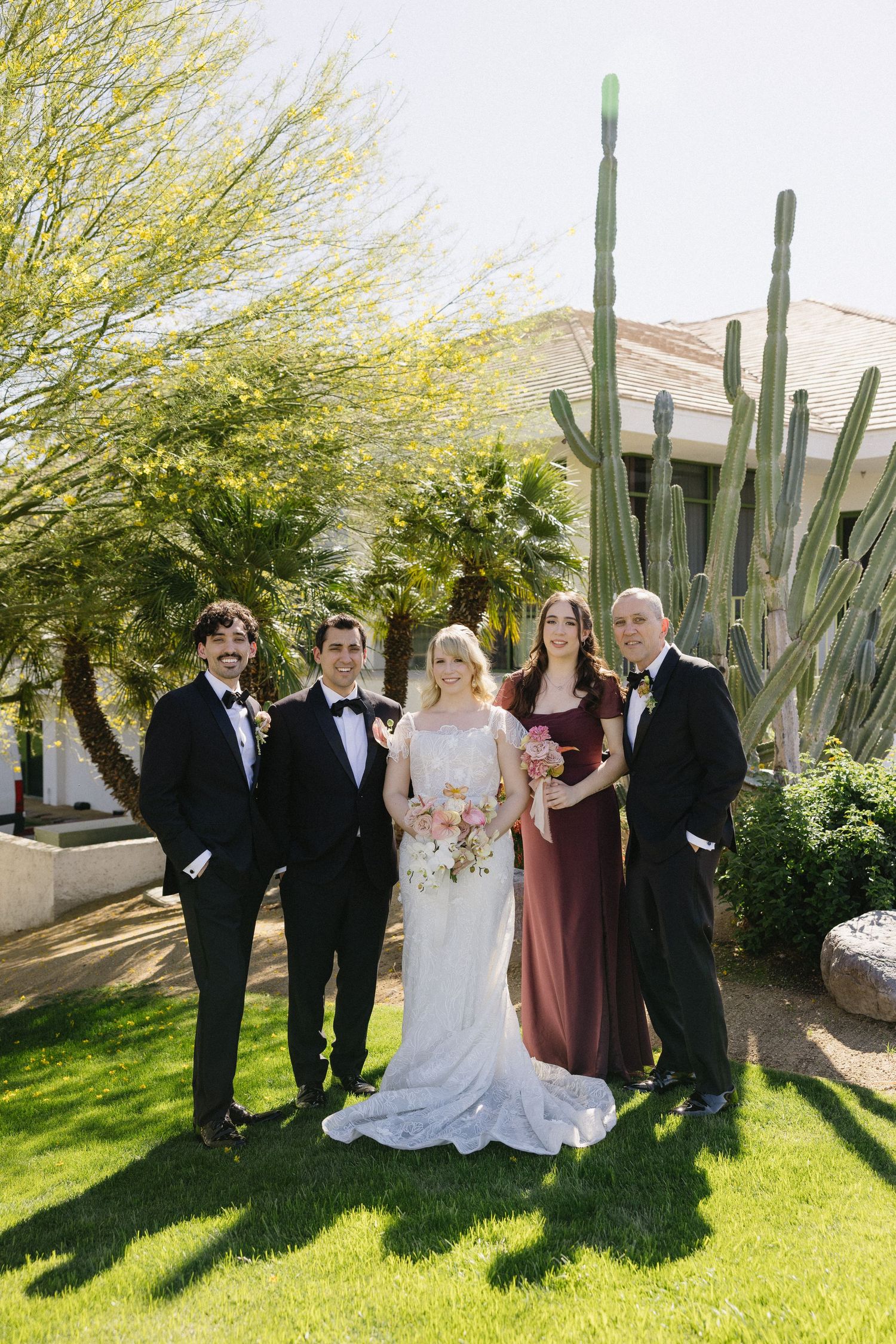 Wedding party of five posing outdoors with tall cactus and green trees, bride in white gown holding pink bouquet.