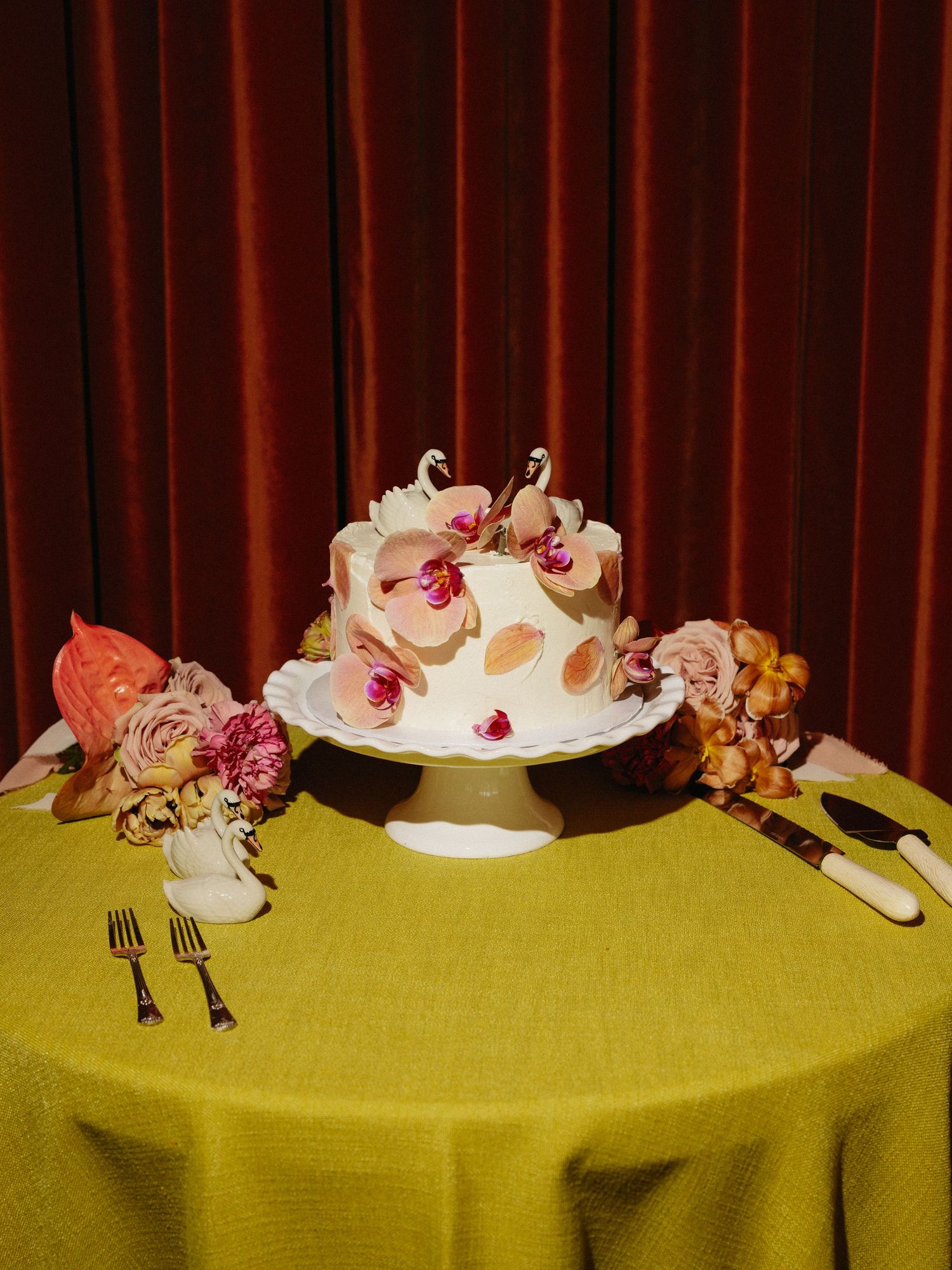 Two-tier white wedding cake with pink orchids and swan toppers on yellow tablecloth with floral arrangements.