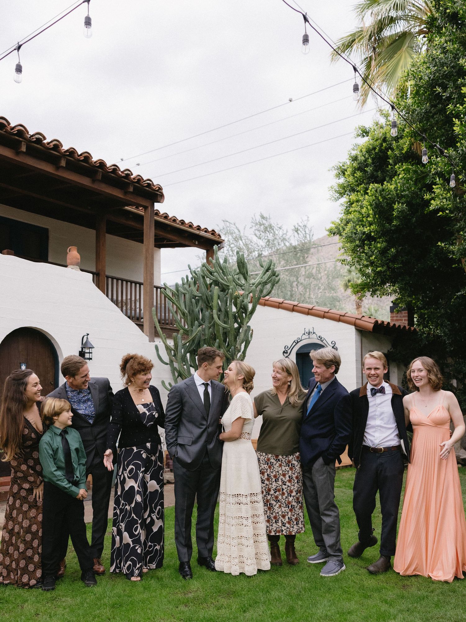 Wedding party group photo outdoors at a Spanish-style venue with string lights, cacti, and lush greenery.