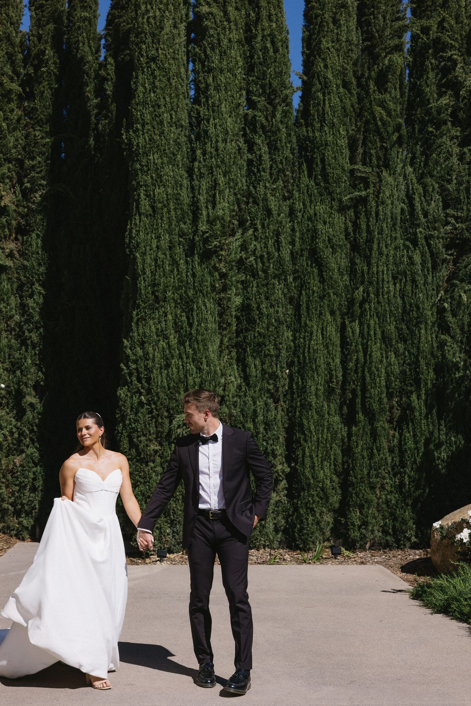 Bride and groom walk hand-in-hand past tall cypress trees on sunny wedding day.