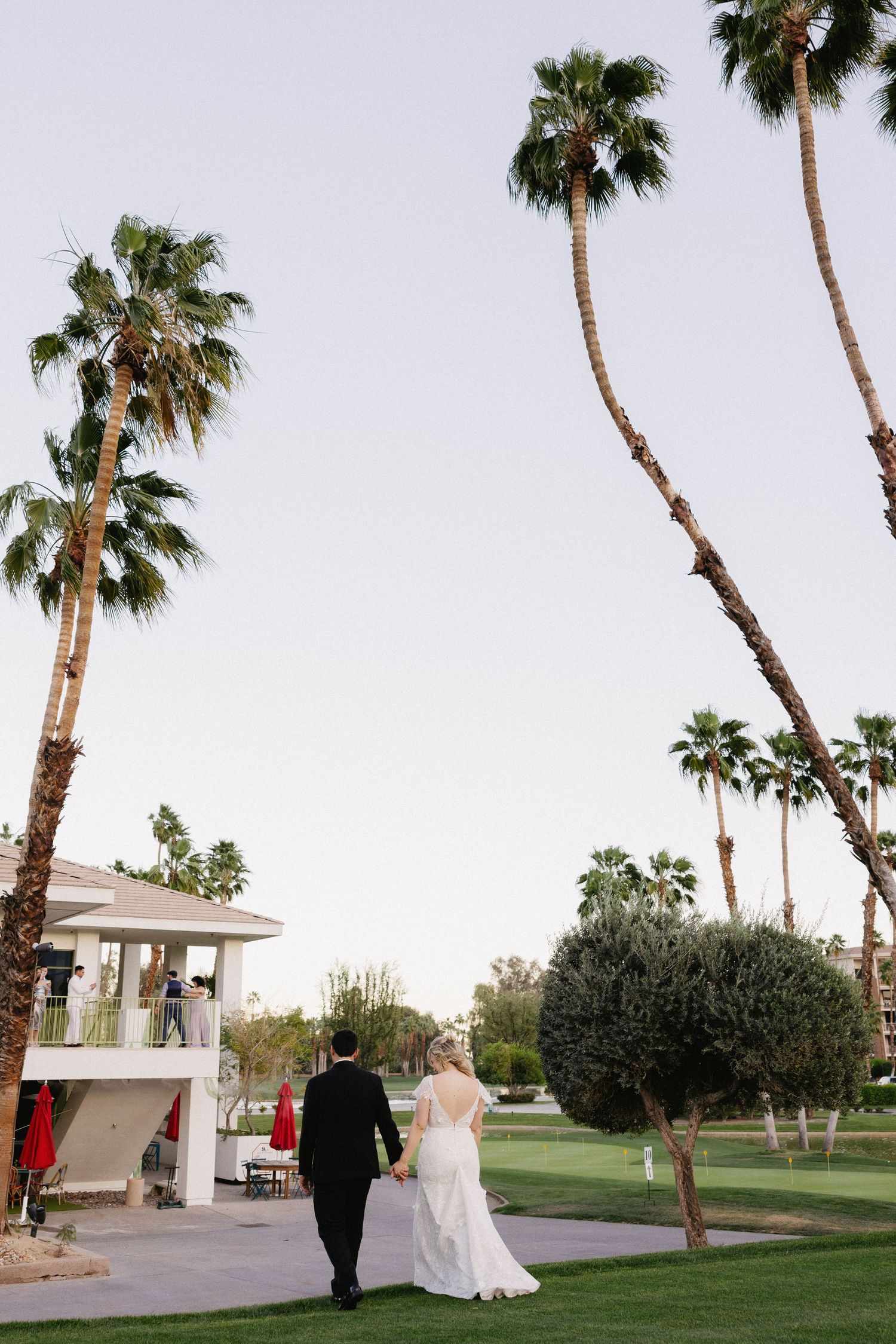 Bride and groom walk hand in hand through lush palm tree-lined grounds at a sunny Palm Springs wedding venue.