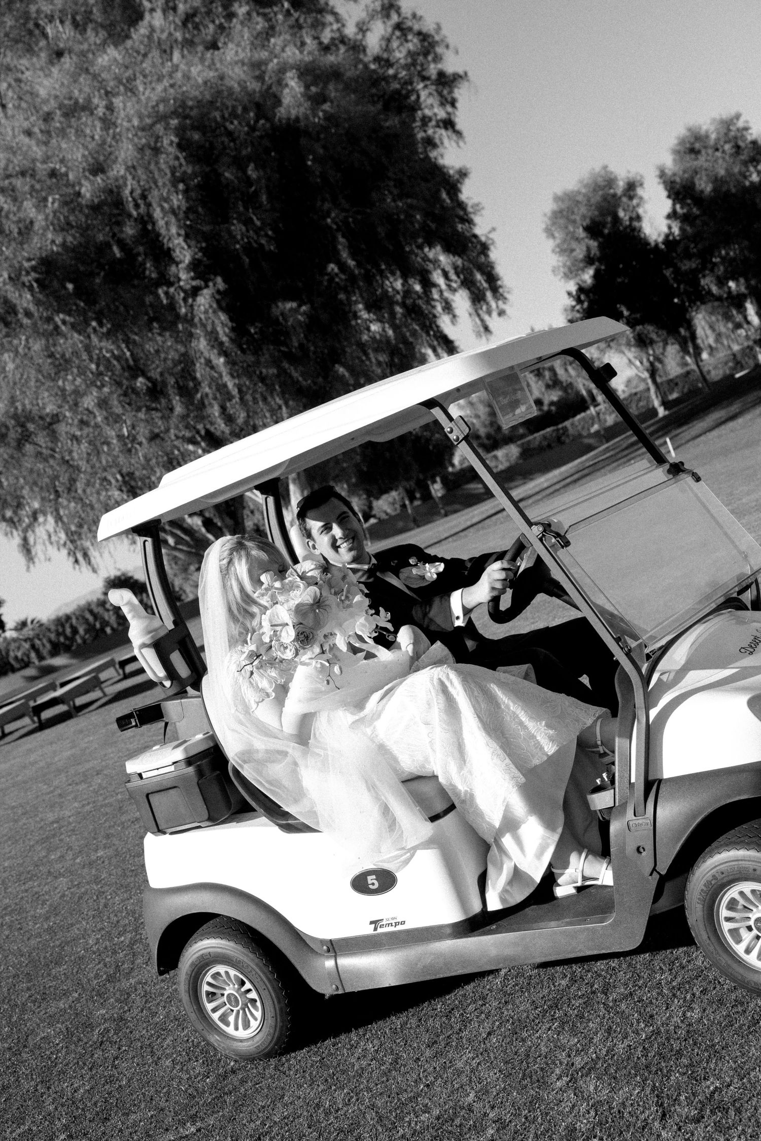 Black and white photo of a bride and groom riding a golf cart on their wedding day, surrounded by trees.