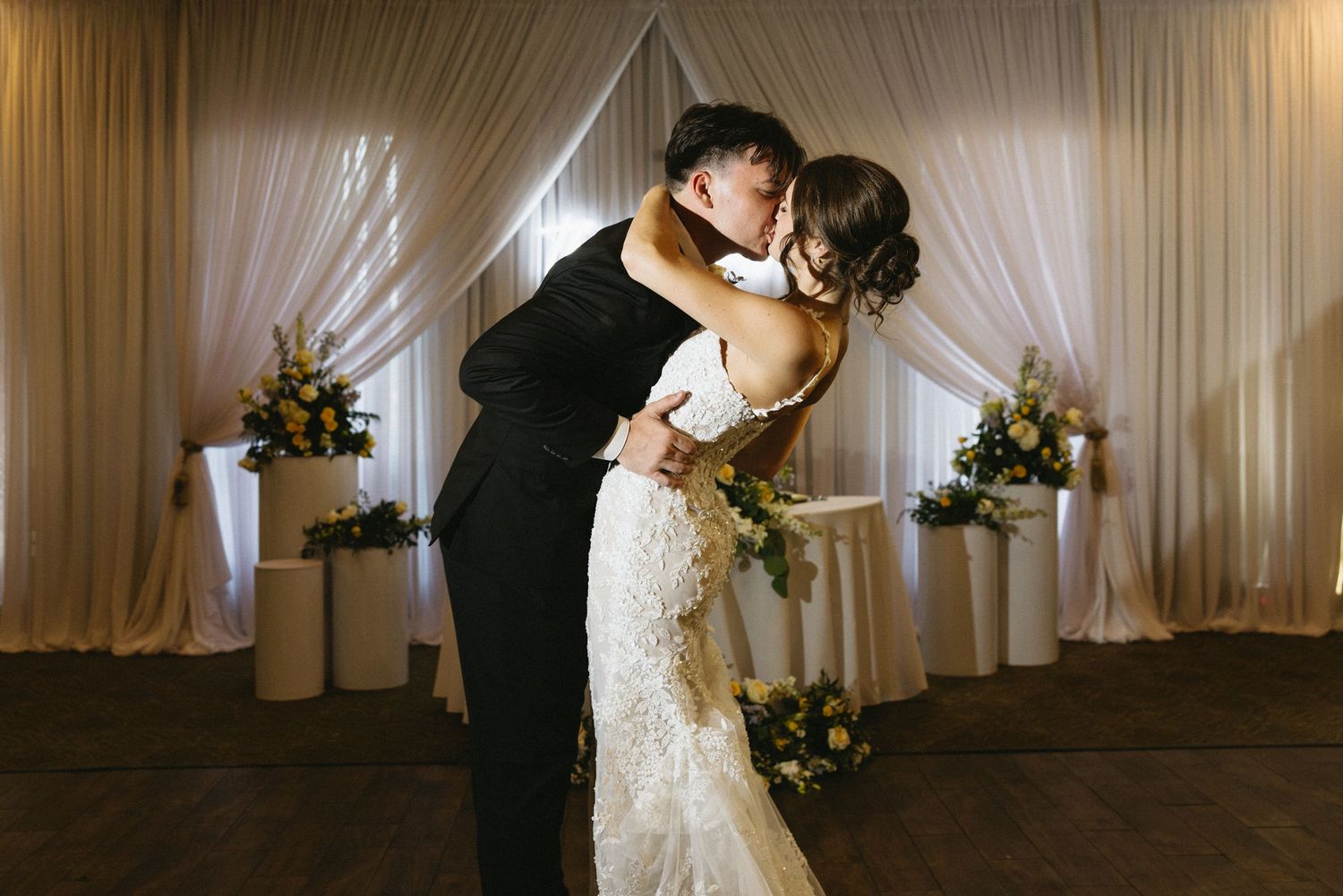 Bride and groom share a romantic first dance kiss in elegant ballroom with white draping and floral arrangements.