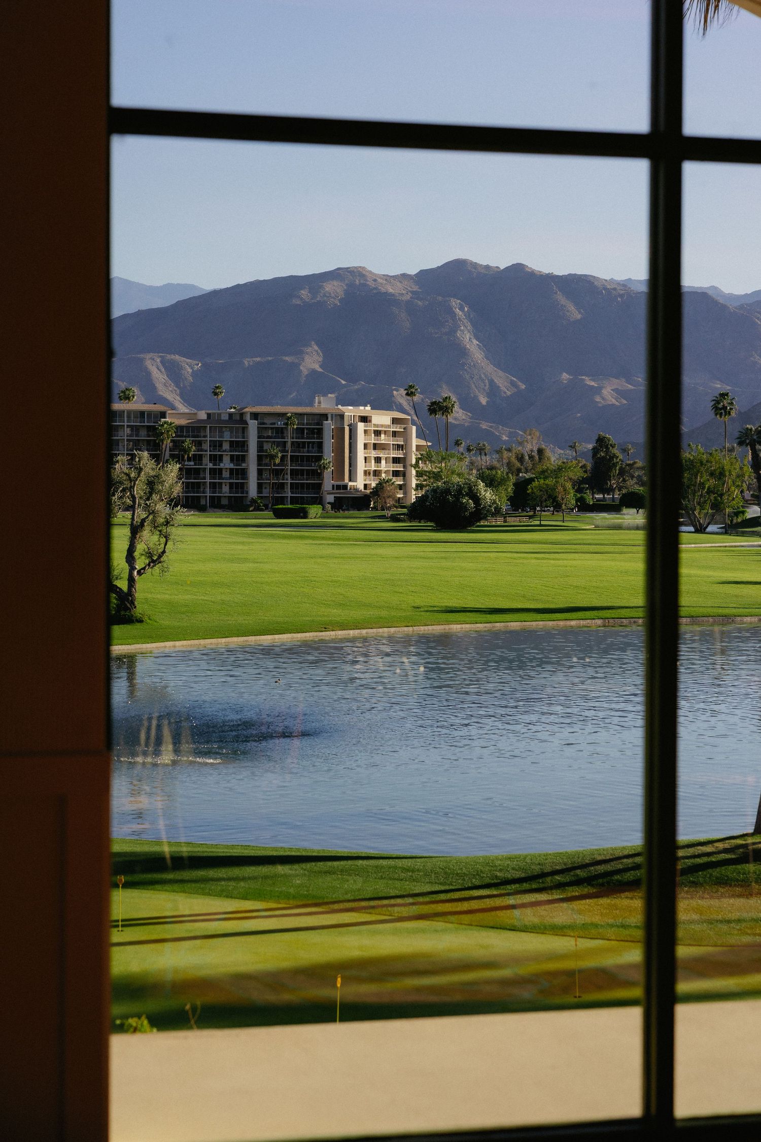 View through window of lush golf course with pond, resort buildings, and mountain backdrop in Palm Springs, California.