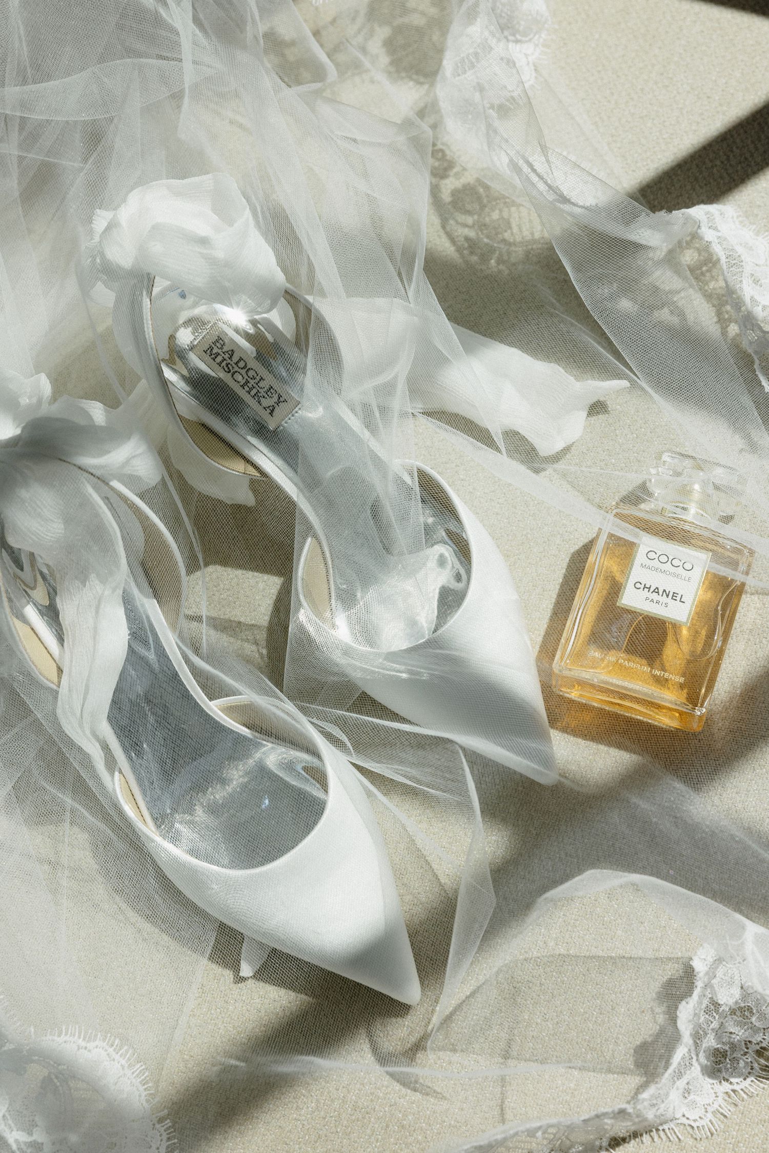 Badgley Mischka white satin bridal heels with ribbon bows beside Chanel Coco perfume bottle on flowing tulle veil.