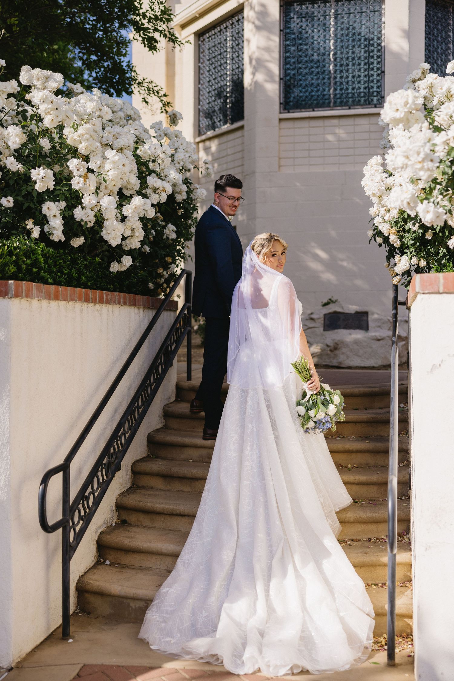Wedding couple walking up white stairs decorated with lush white floral arrangements.