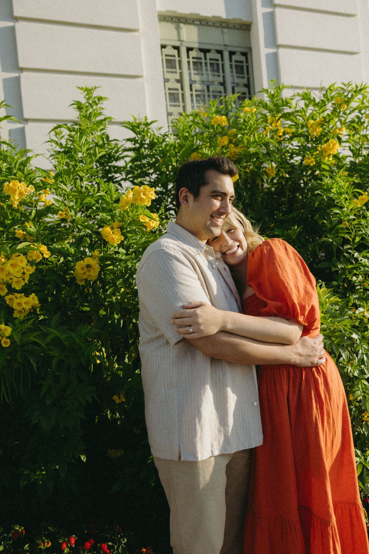 Couple embracing in front of yellow flowers and white building wall.
