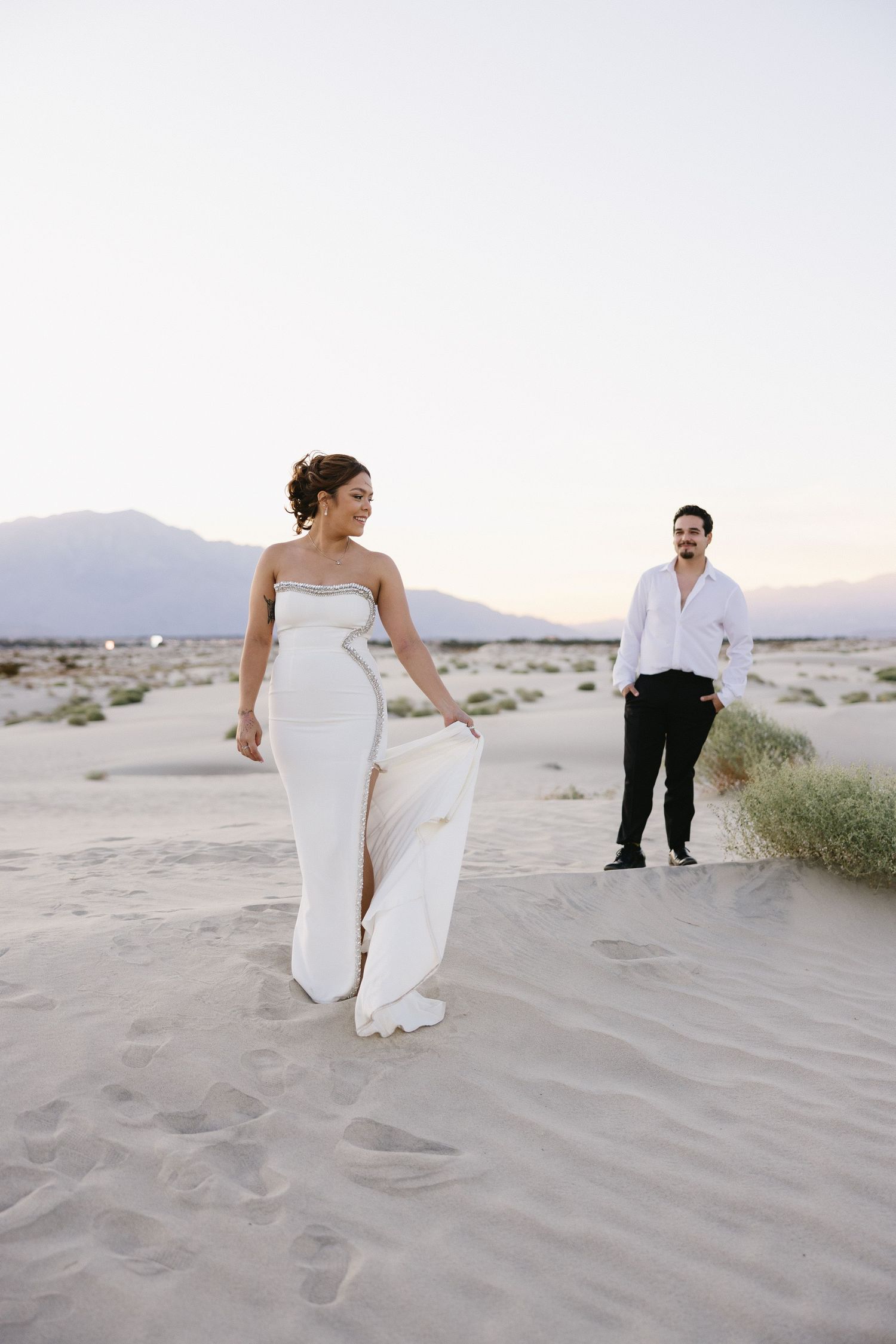 Elegant wedding photo of couple posing on sand dunes with mountains in background at dusk.