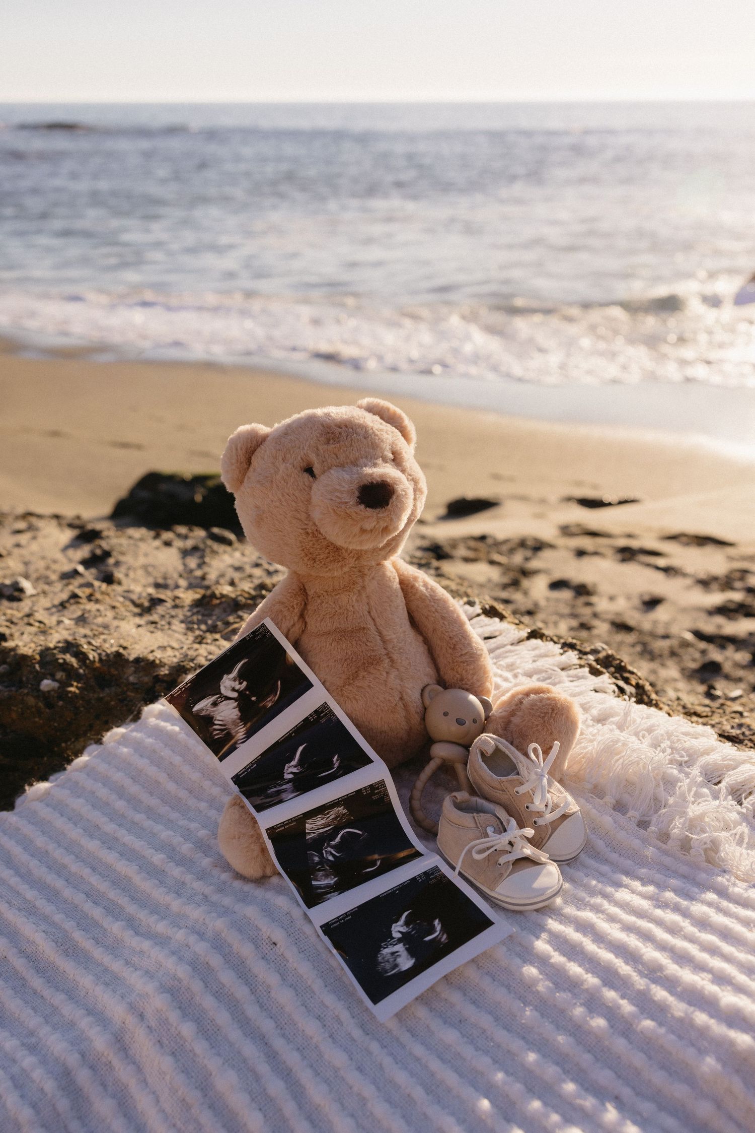 Teddy bear on beach blanket with ultrasound photos in golden sunlight.