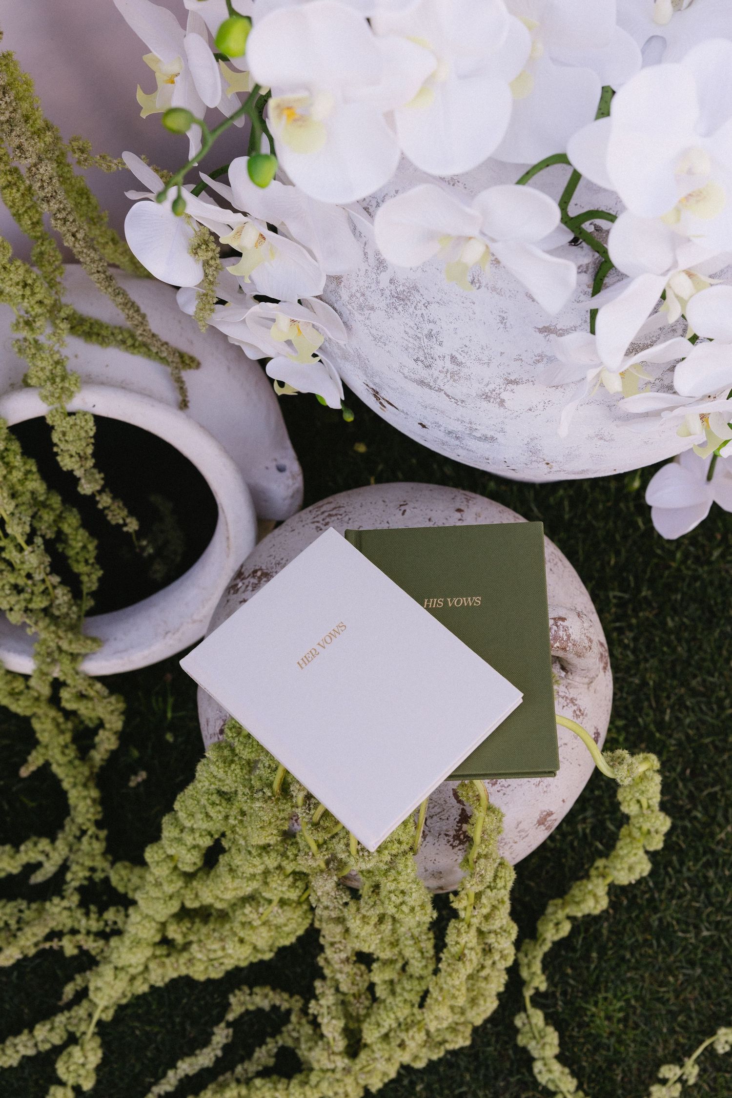 Elegant wedding vow books on a stone pedestal surrounded by white orchids and green moss in a garden setting.