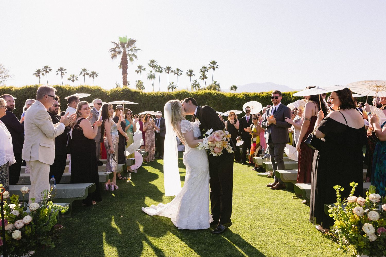 Bride and groom share first kiss at outdoor wedding ceremony surrounded by guests on sunny lawn with palm trees.