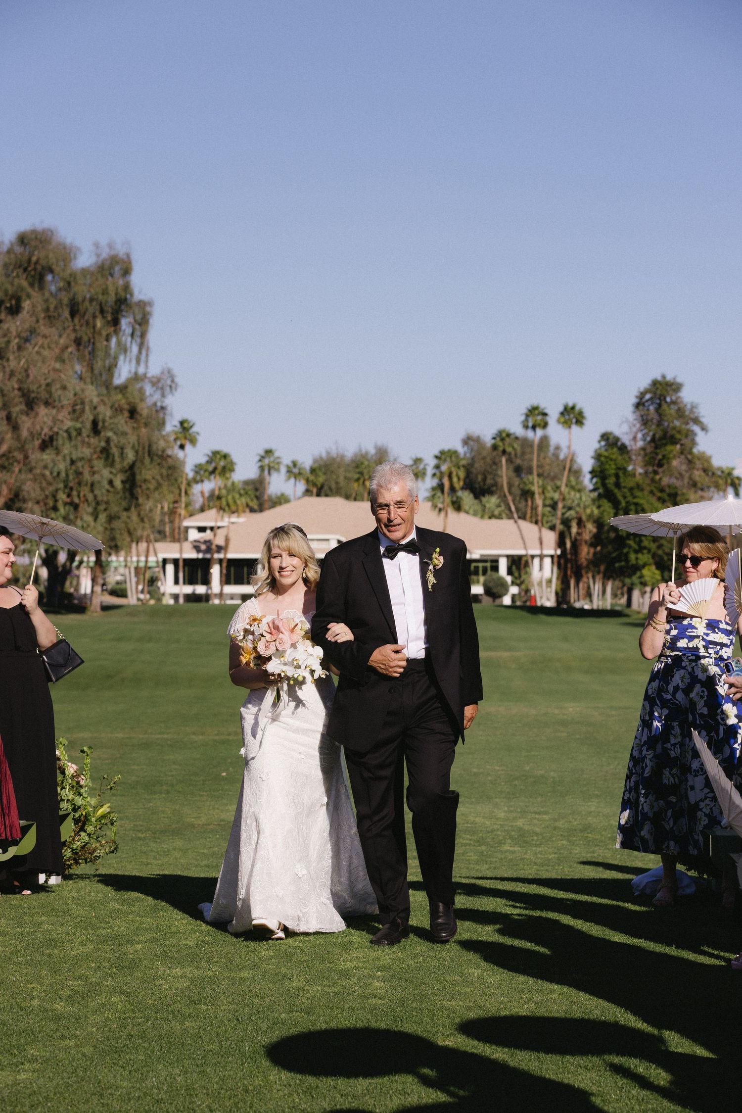 Bride in white gown walks down outdoor aisle with older man in tuxedo at sunny garden wedding ceremony.