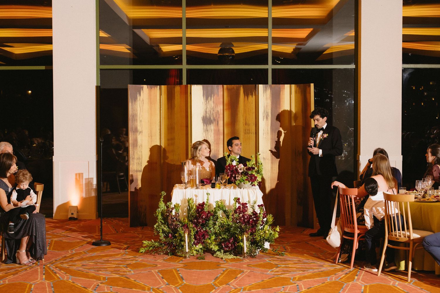 Elegant wedding reception with couple at sweetheart table adorned with lush floral arrangements in a warmly lit venue.